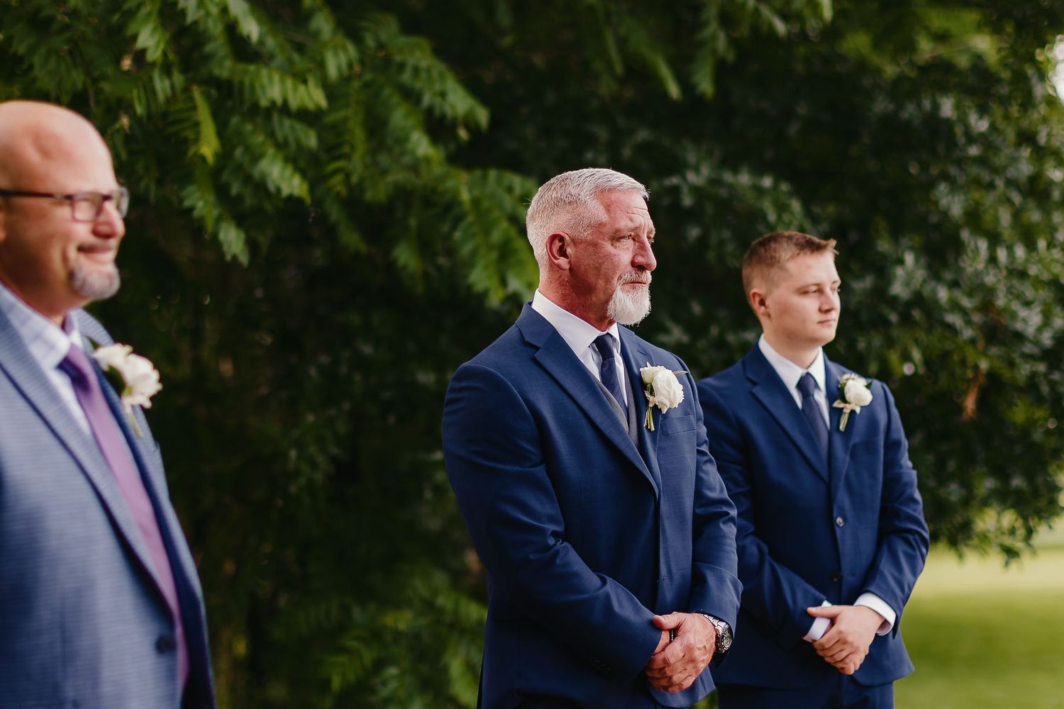 Three men in blue suits standing during outdoor wedding ceremony.