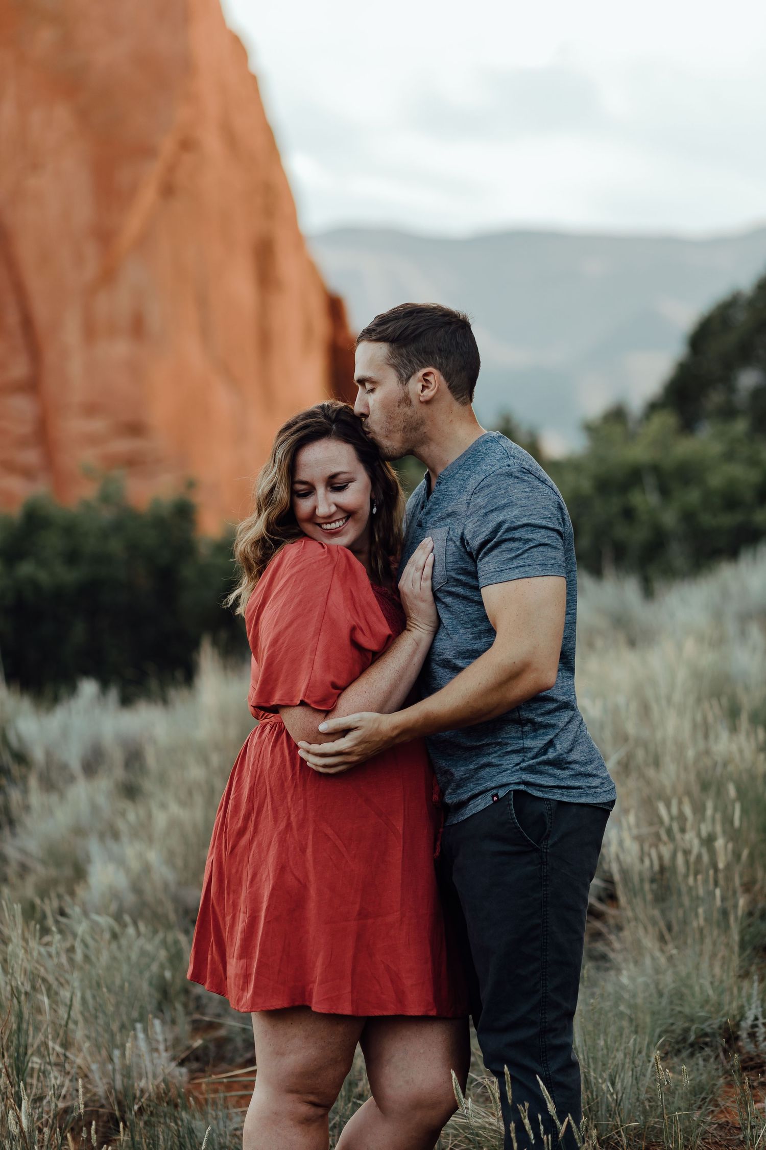 A romantic couple embraces against a backdrop of red rocks and mountains in an outdoor setting.
