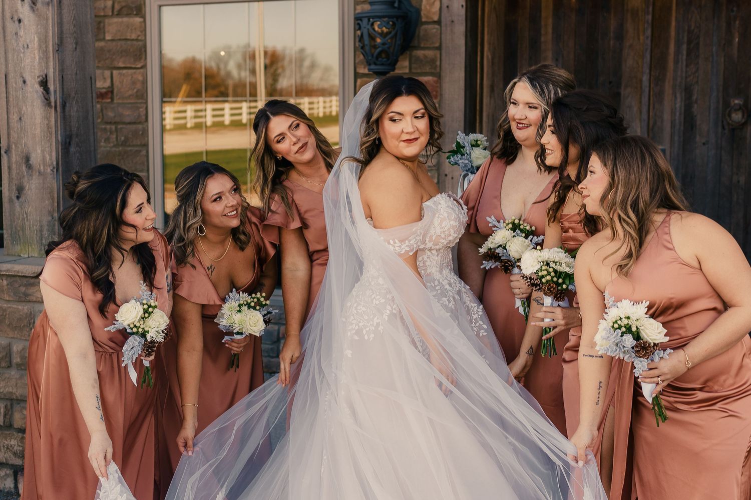 Wedding party poses together in dusty rose bridesmaid dresses and a white bridal gown against brick wall.