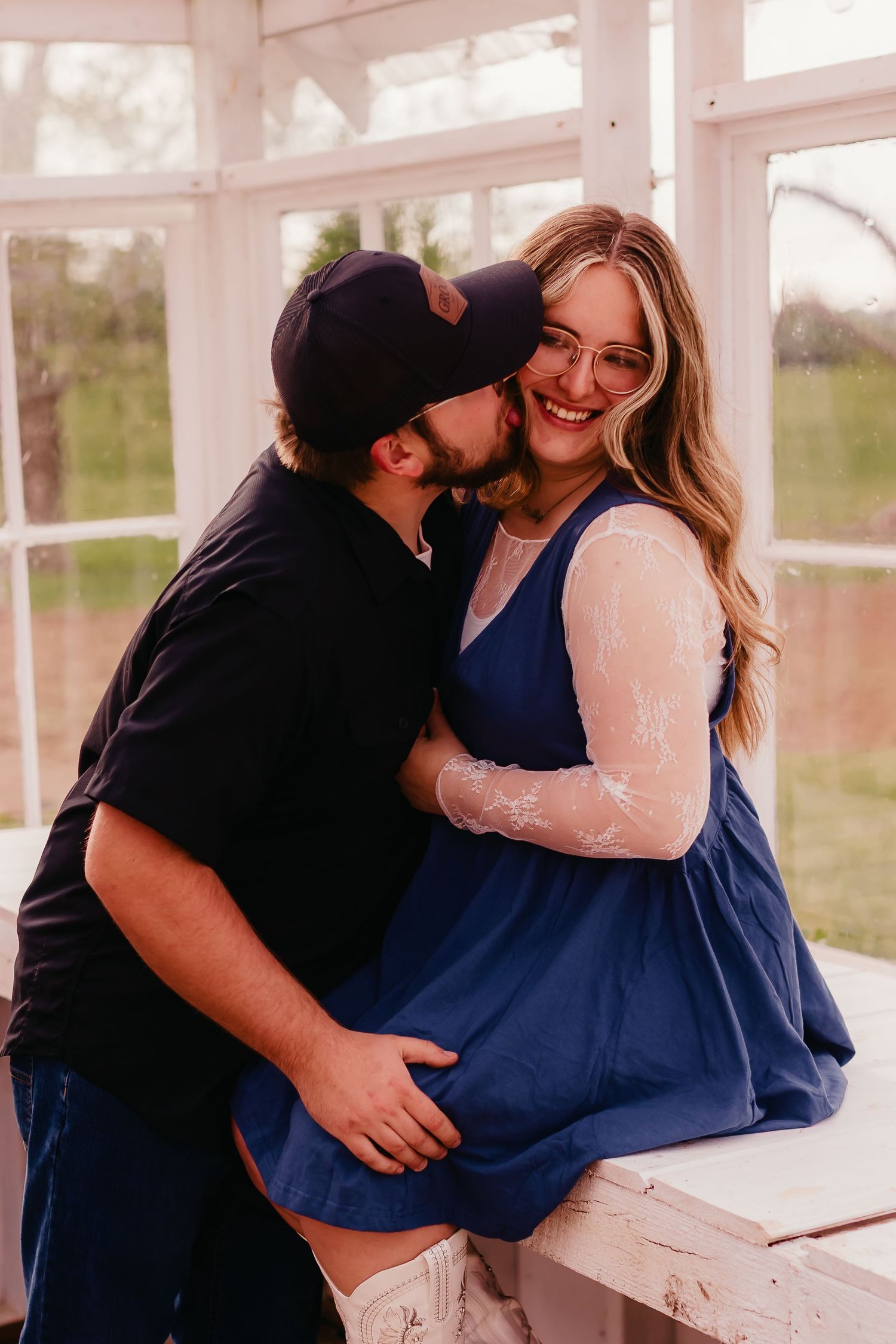 A couple shares a tender moment as a man kisses a smiling pregnant woman in a blue dress near bright windows.
