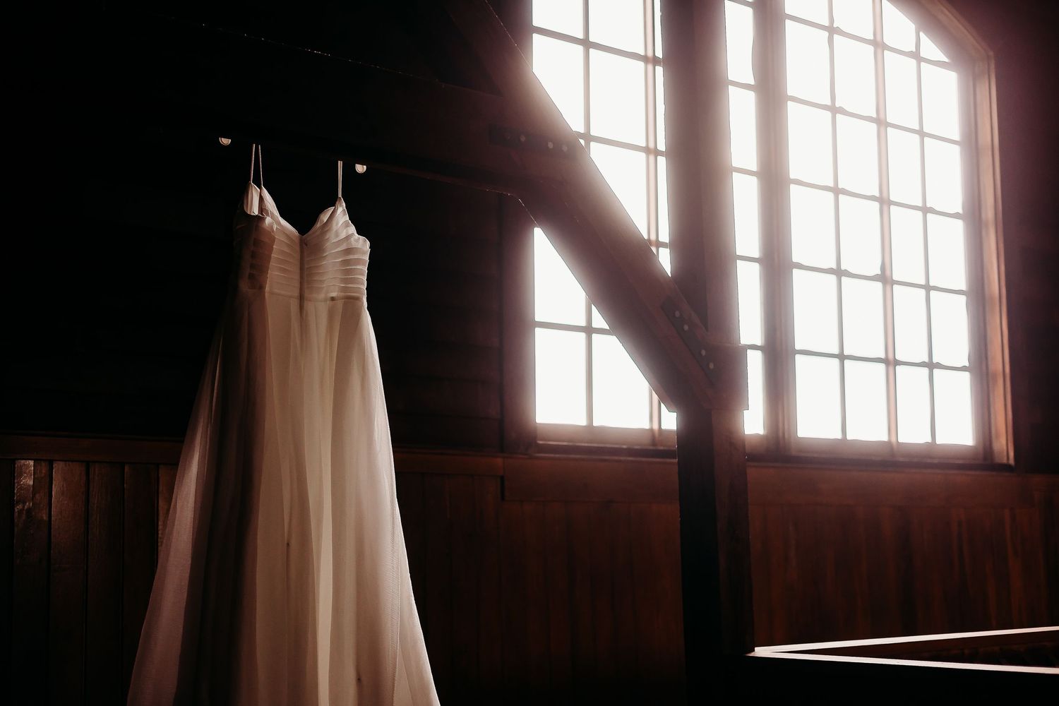 White wedding dress hanging in front of sunlit windows in rustic wooden room.