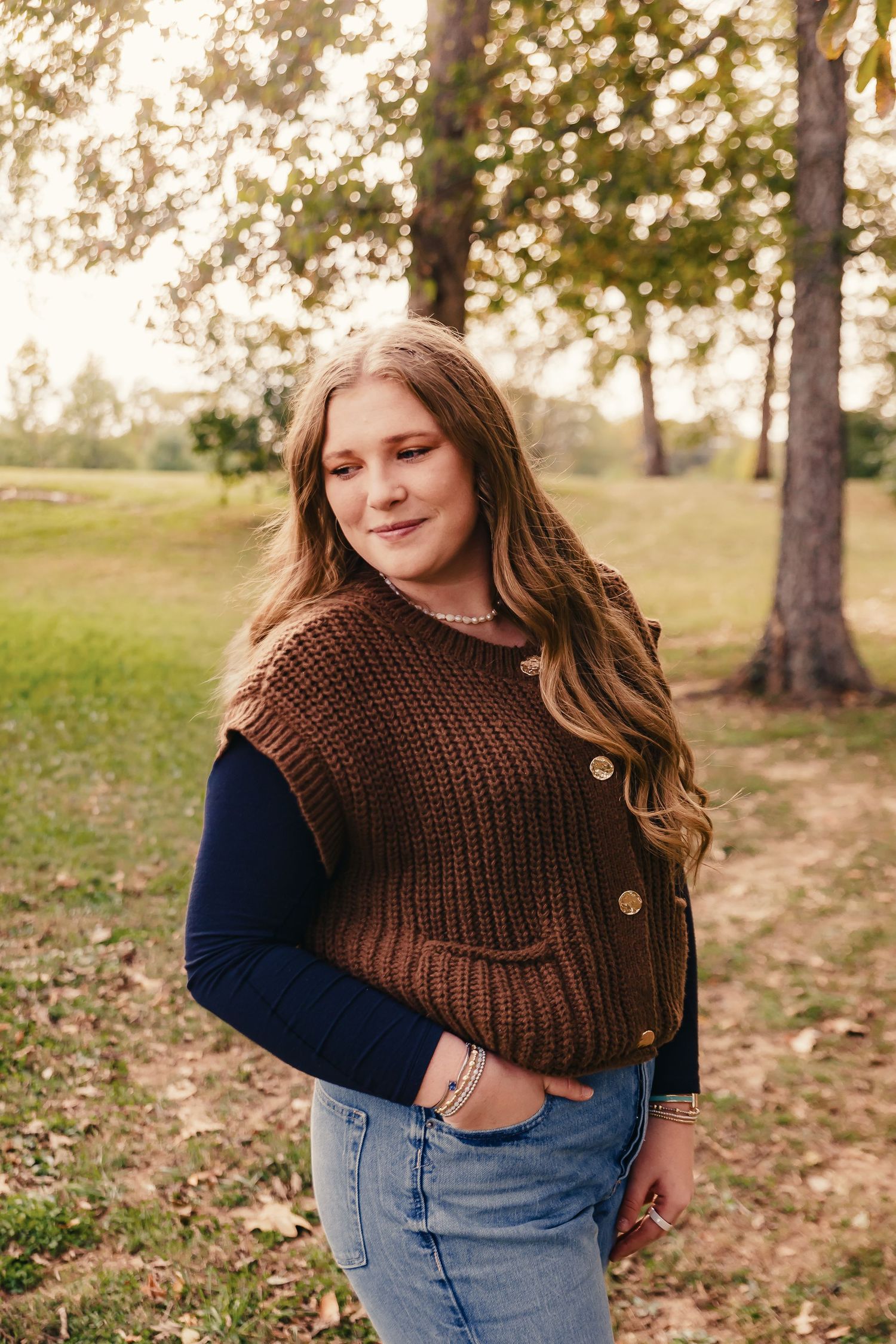 A person in a brown knit sweater poses outdoors against an autumn landscape background.