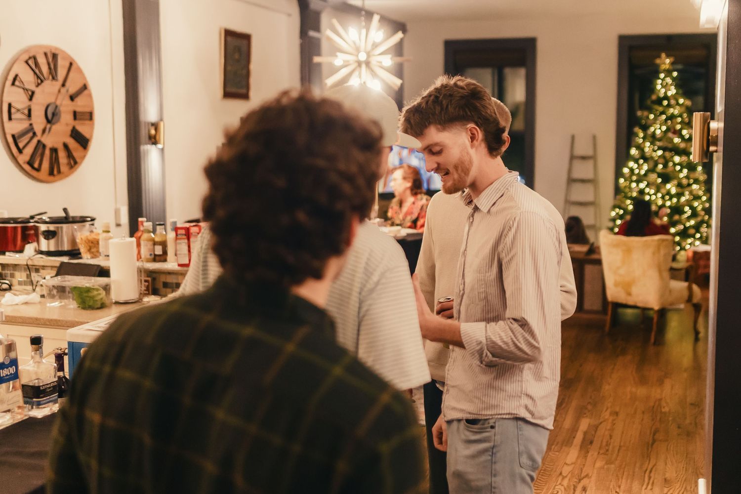 A group of people gather around a dining table during a holiday celebration with a Christmas tree in the background.