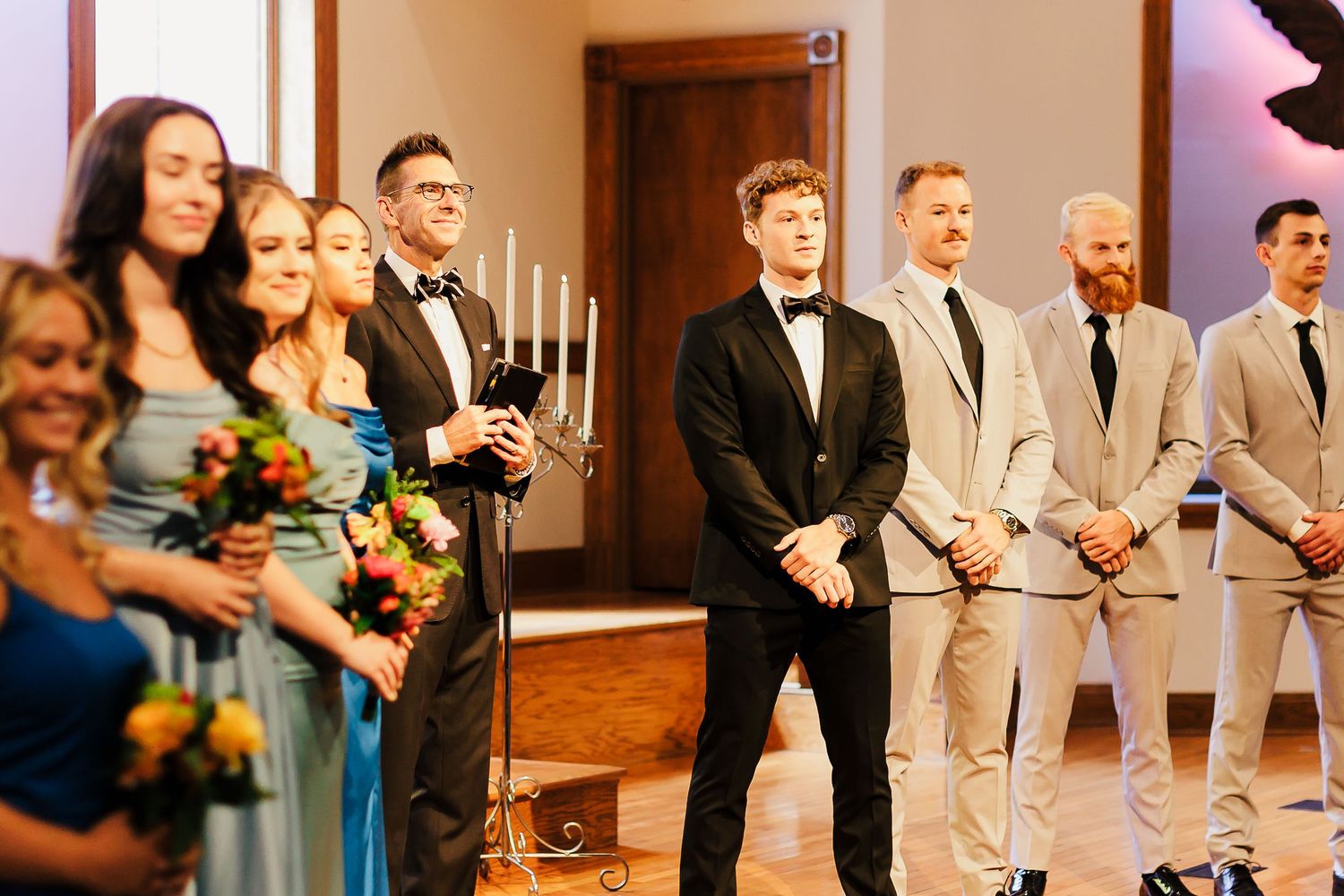 Wedding party poses together in formal attire at indoor ceremony venue.