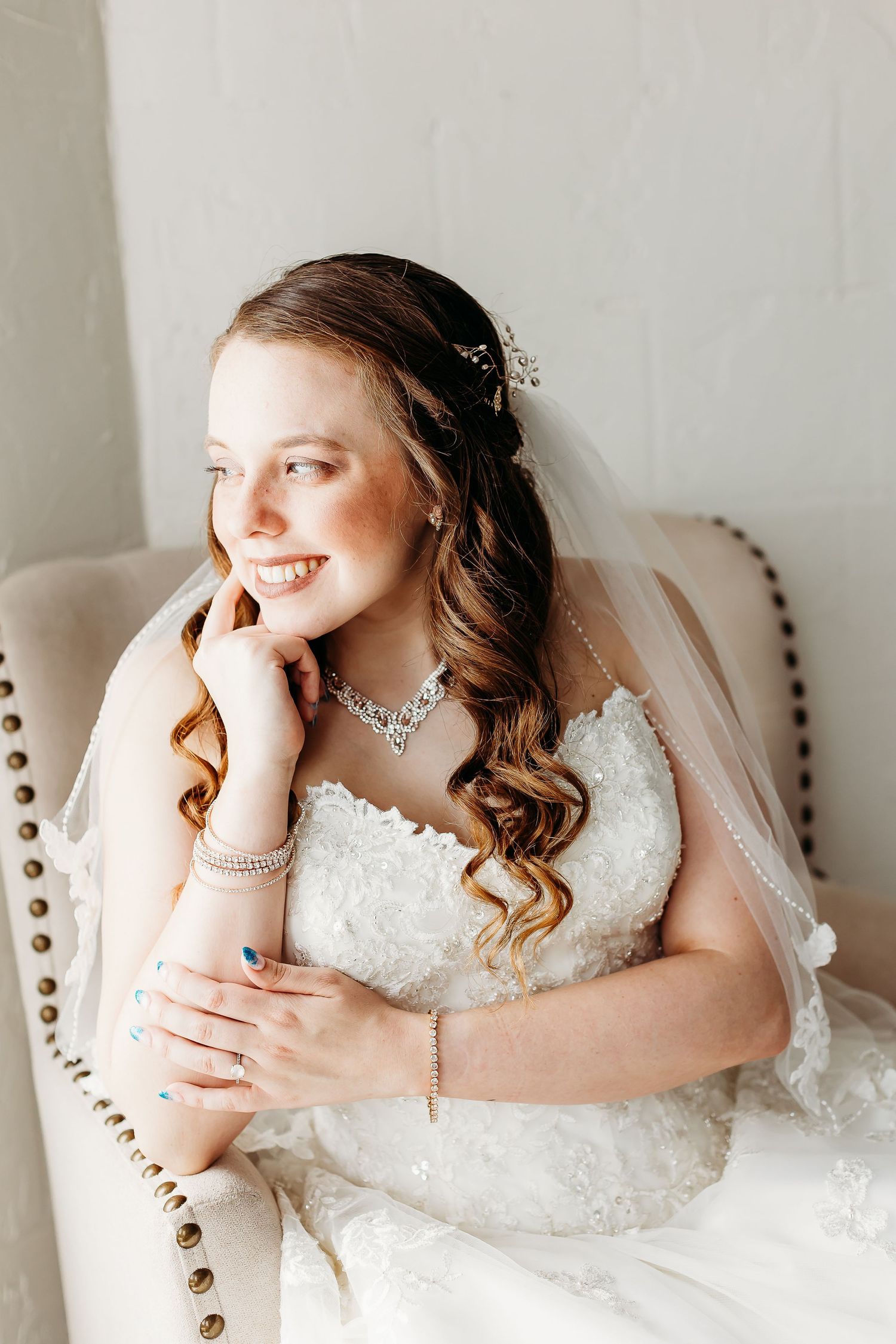 Elegant bridal portrait on tufted chair showing jewelry and lace dress details.