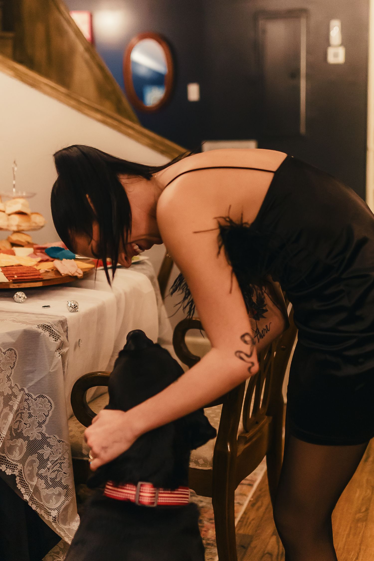 Woman in black dress bends over while playing beer pong at a party on wooden table.