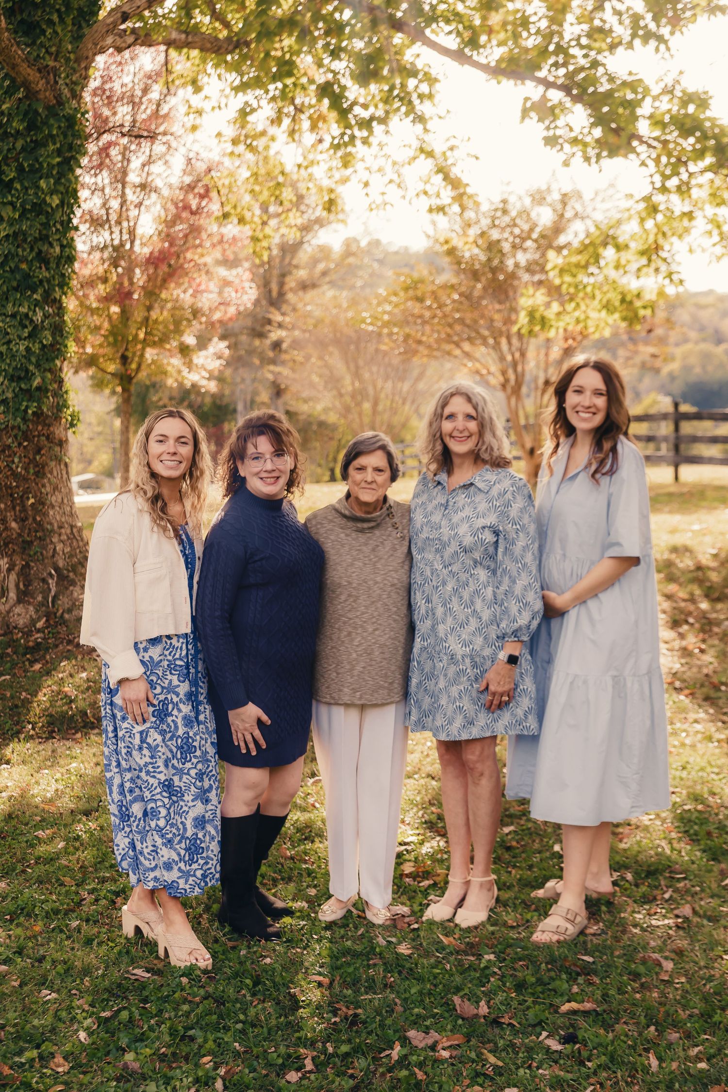 Five women in coordinated outfits pose together outdoors during golden hour with a rustic fence backdrop.