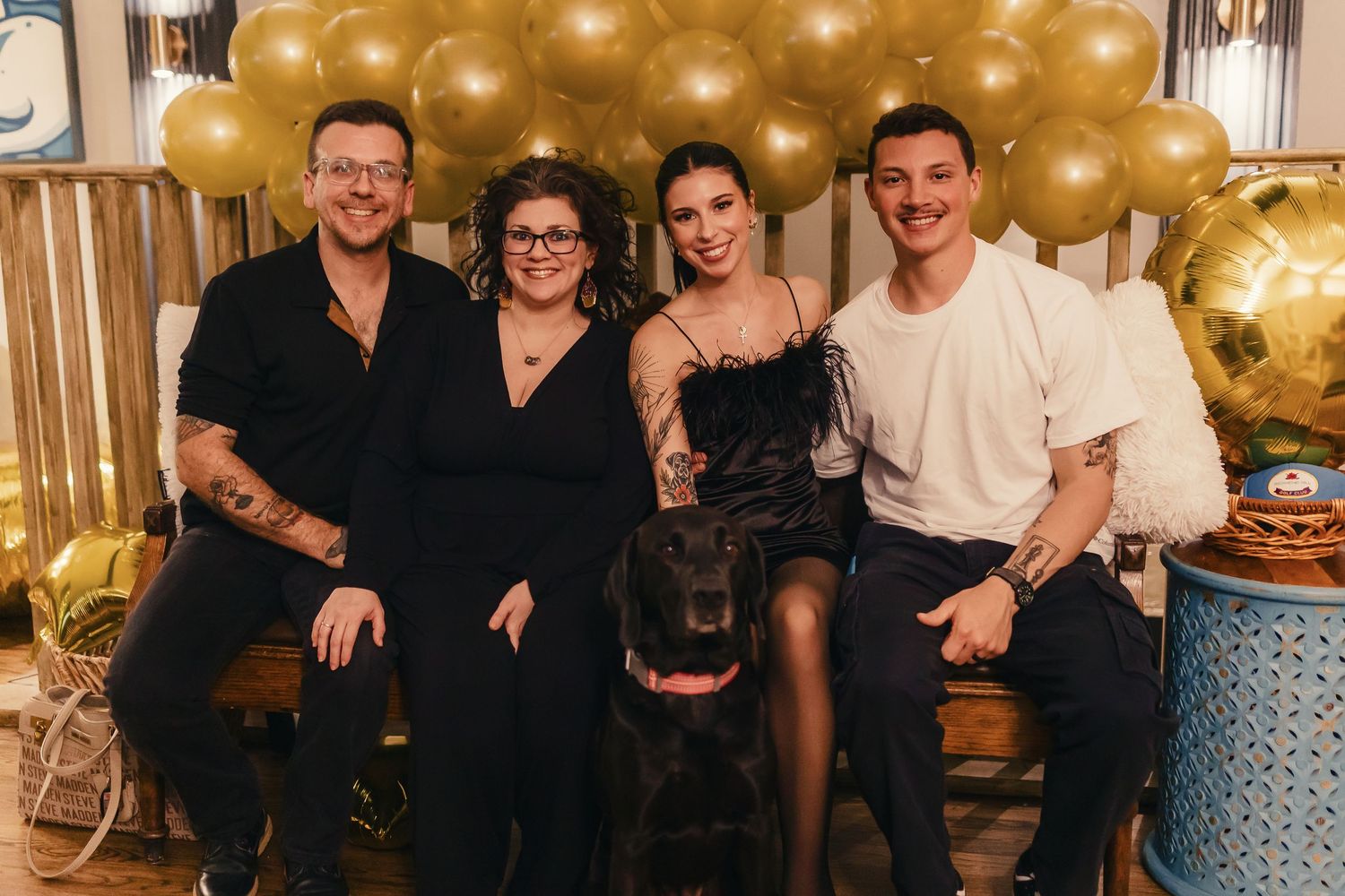 A group poses together at a celebration with gold balloon decorations and a black dog in the foreground.