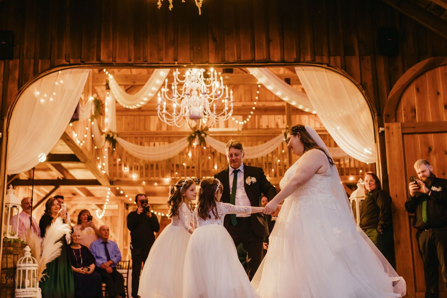 Wedding reception venue decorated with chandeliers and white drapery in rustic barn setting.