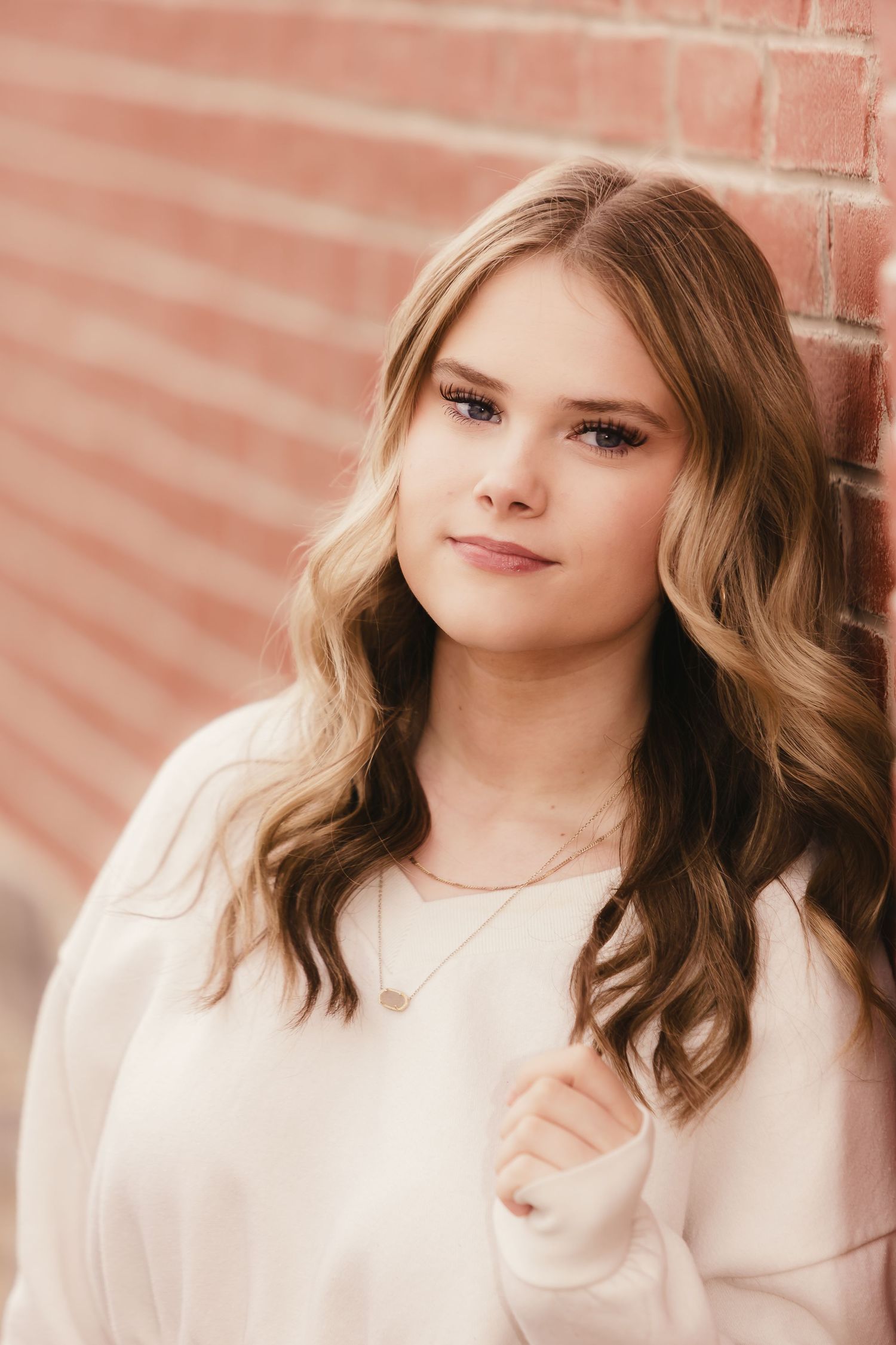 Young woman with wavy blonde hair in white sweater leaning against a brick wall, wearing a delicate necklace.