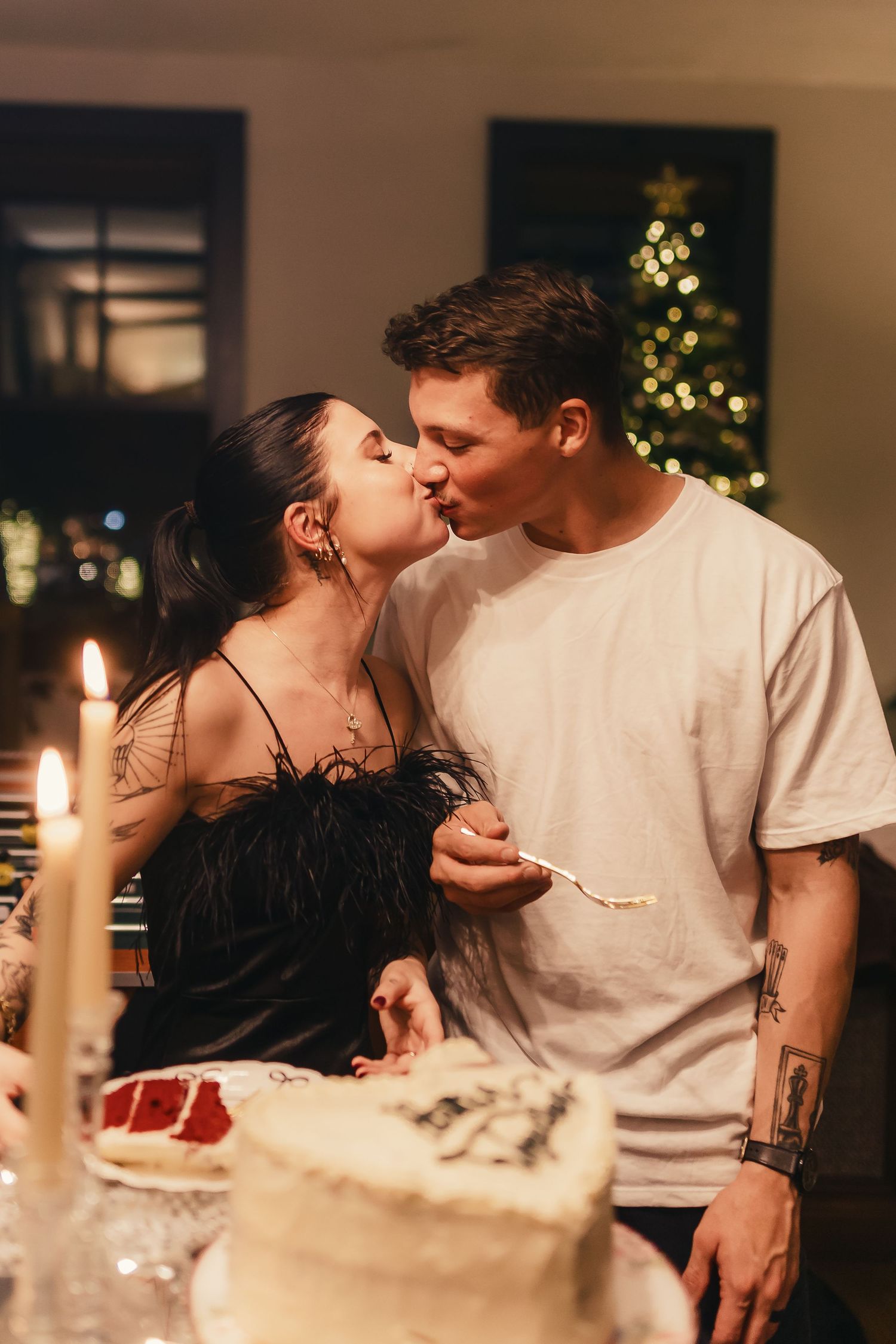 A couple shares an intimate moment at a candlelit dinner table with a Christmas tree glowing in the background.