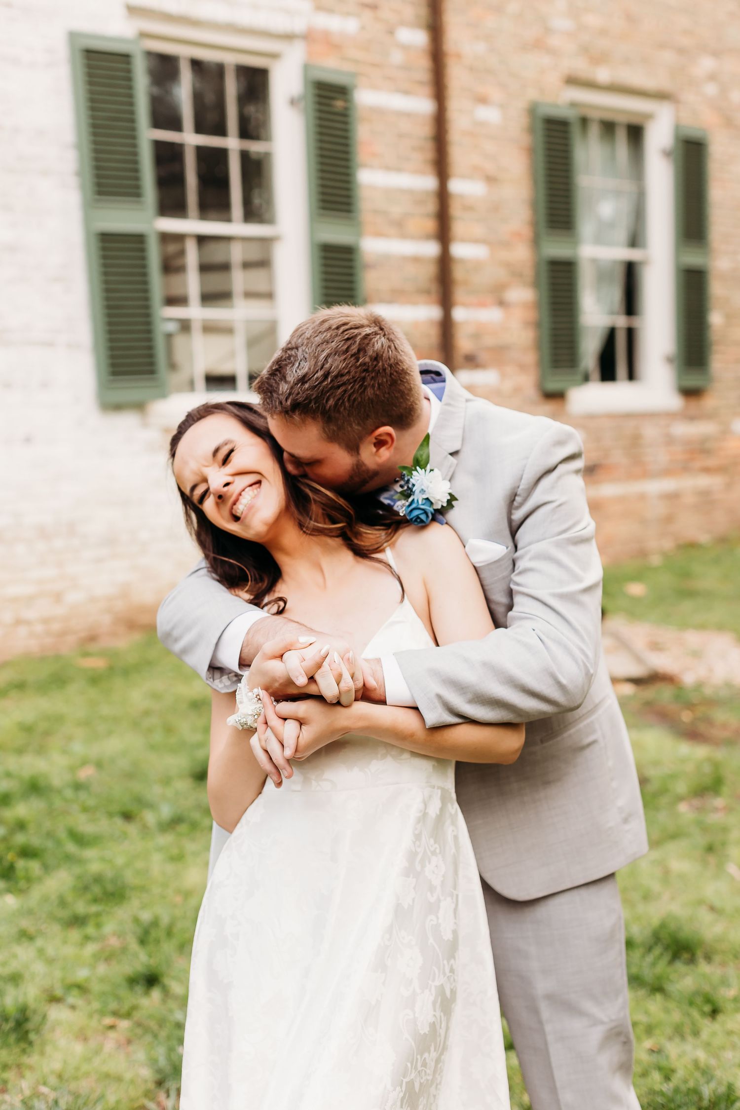 Romantic wedding couple embracing against a brick building with green shutters.