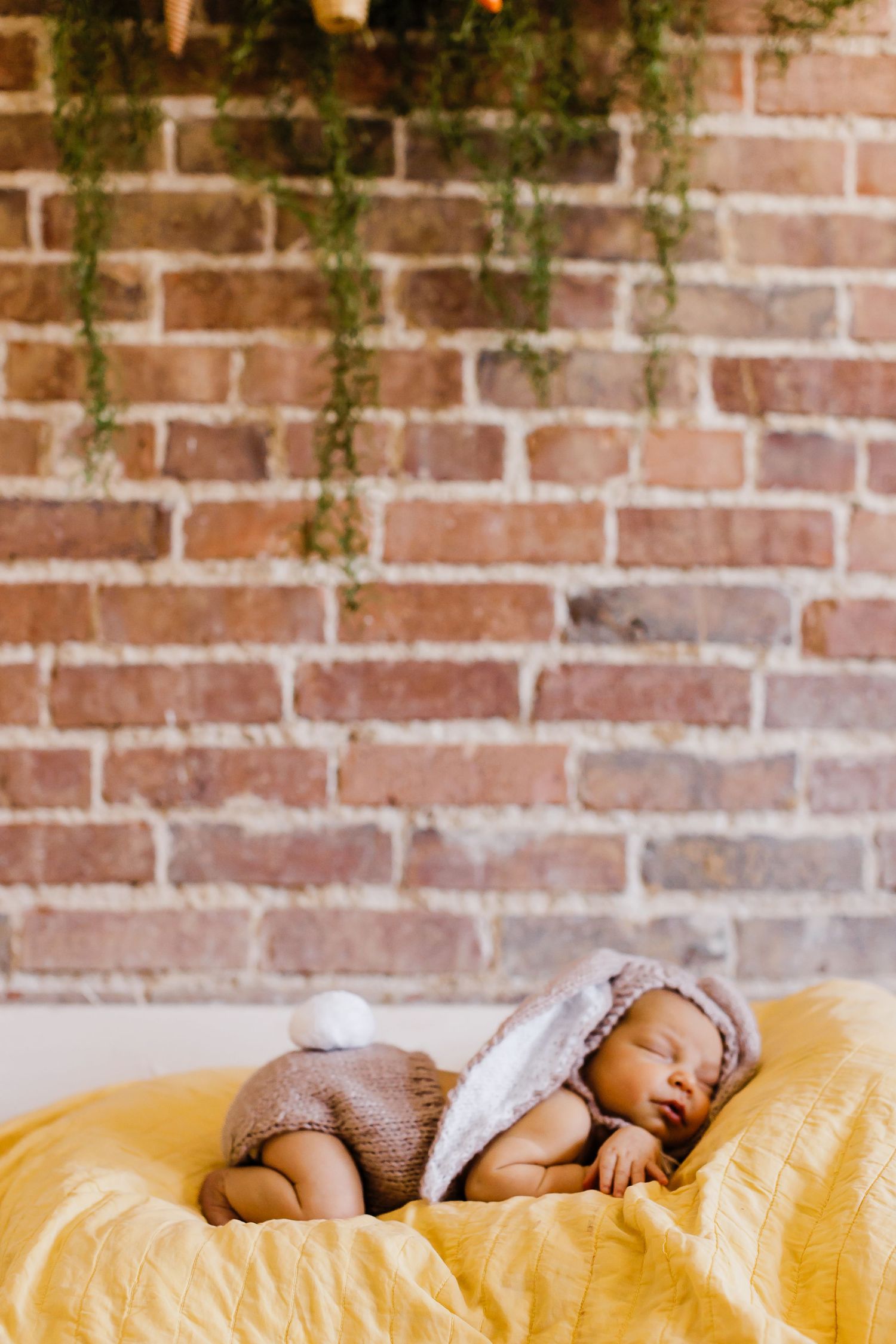 A sleeping infant wrapped in gray swaddle lying on yellow bedding against a brick wall.