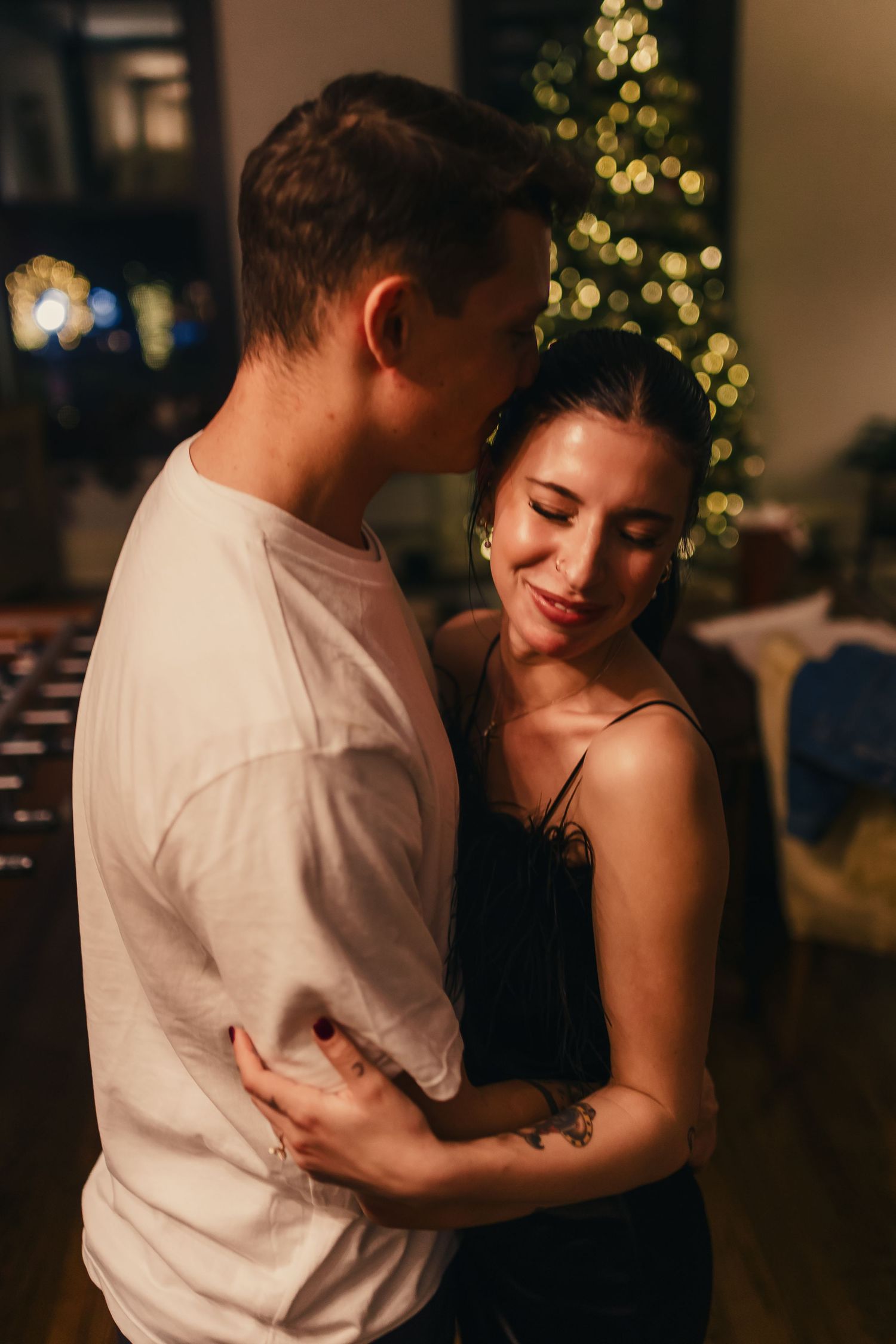 A couple shares an intimate moment while dancing in front of a Christmas tree with soft bokeh lights.