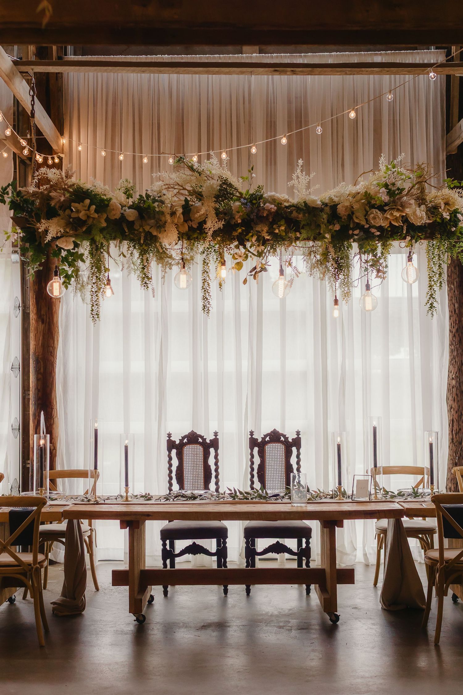 Rustic wedding reception venue with white drapes, string lights, and floral garlands decorating the ceiling beams.