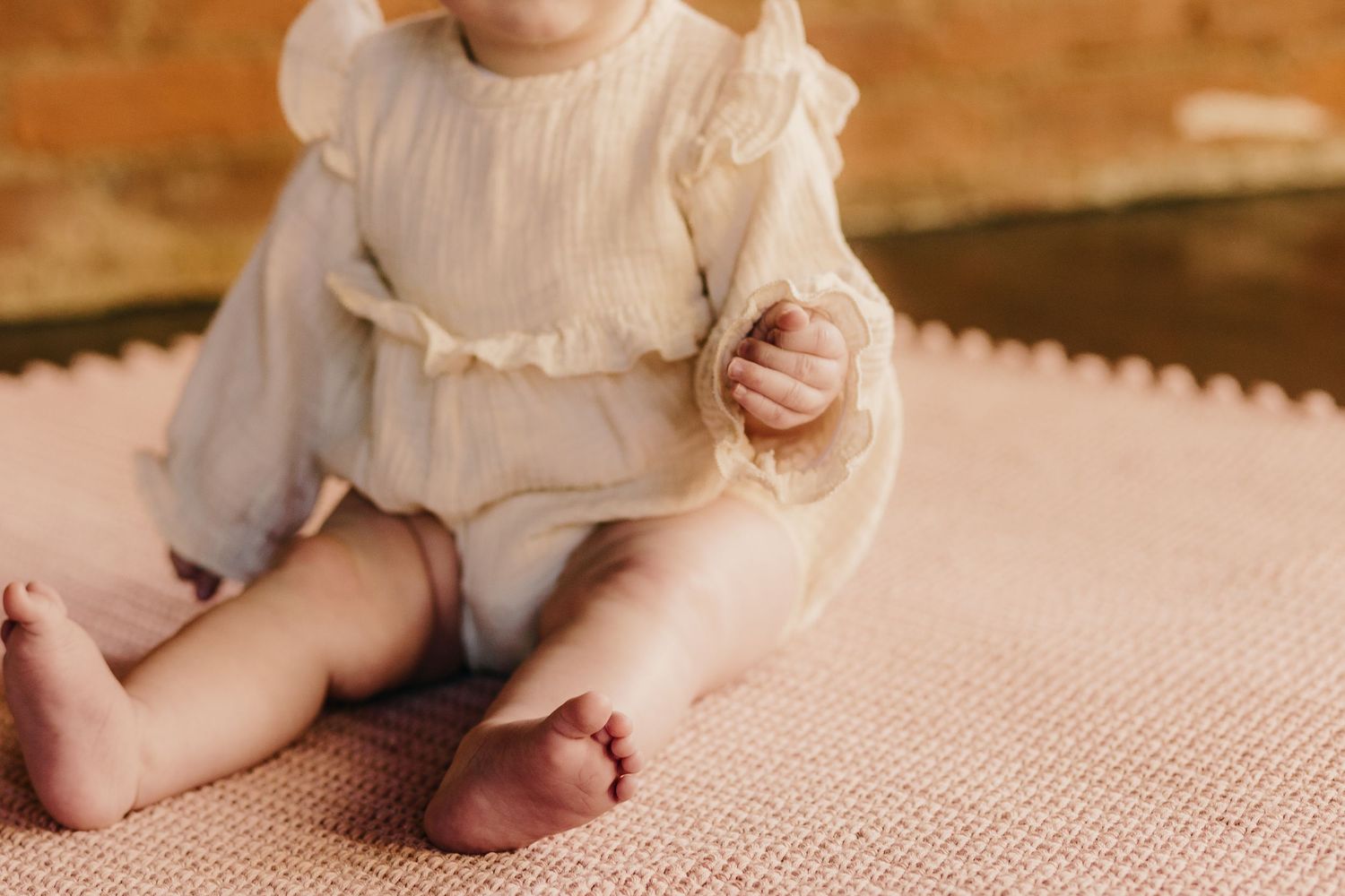 Infant wearing a light-colored dress sits on pink textured fabric.