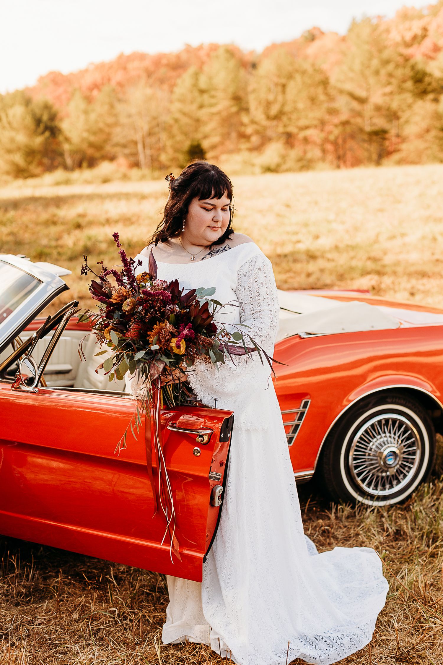 Bride in vintage lace dress holds fall bouquet next to classic red convertible car.