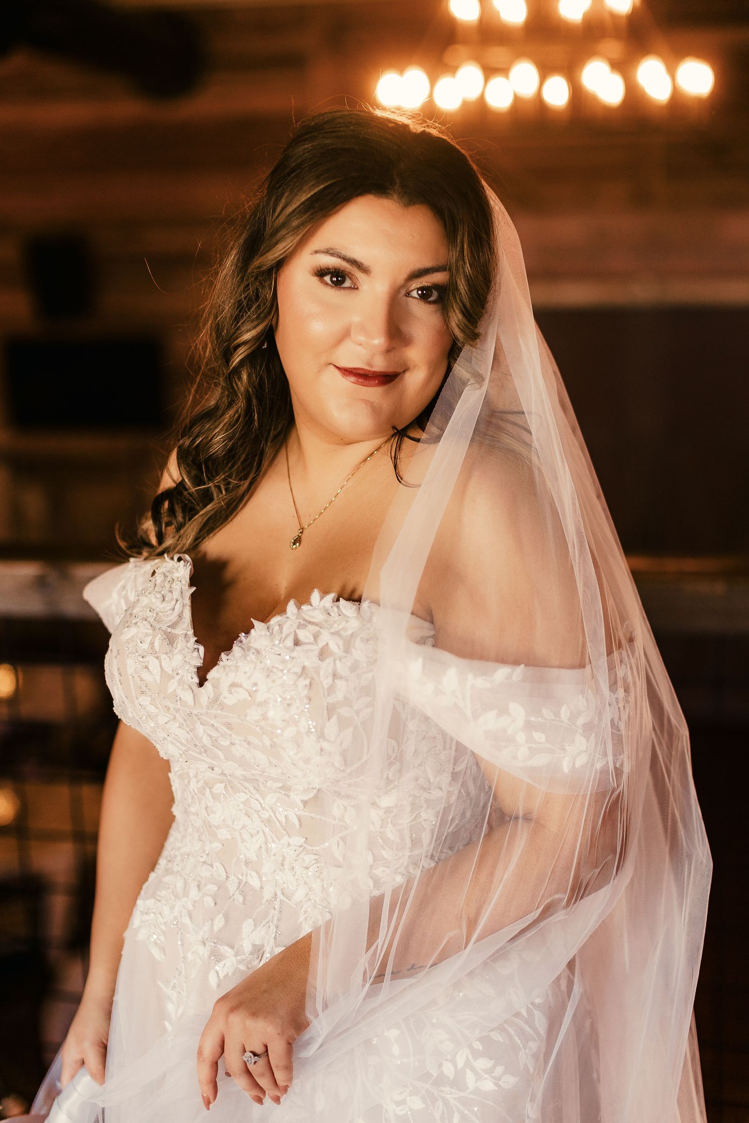 Bride in detailed lace wedding dress with flowing veil poses in warm indoor lighting.