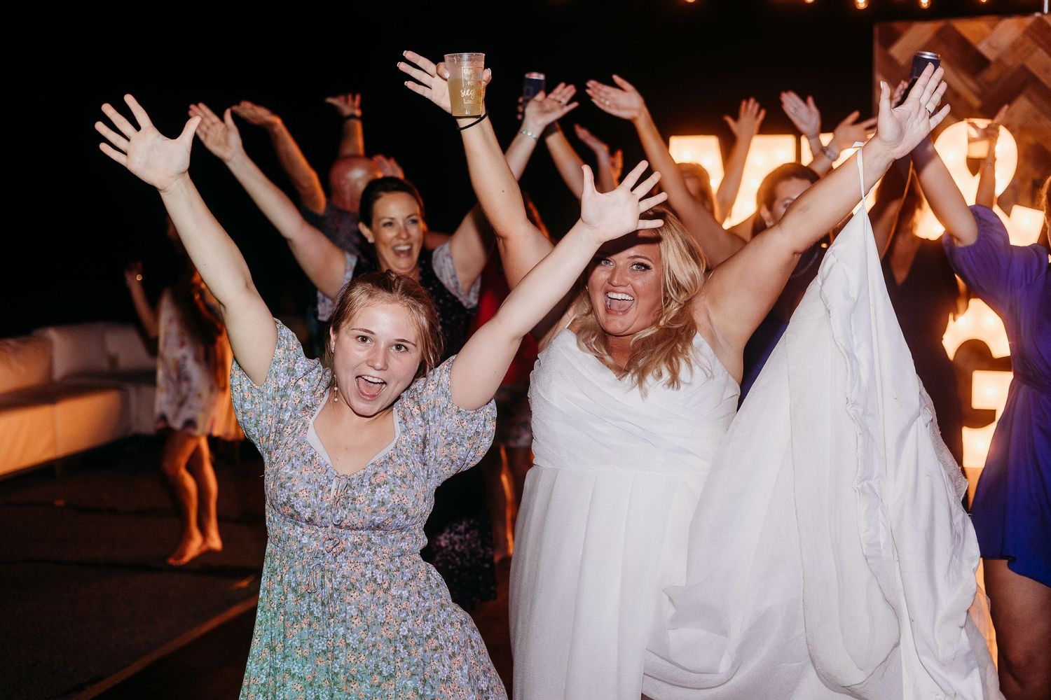 Wedding guests dancing and celebrating with raised arms on a dimly lit dance floor.