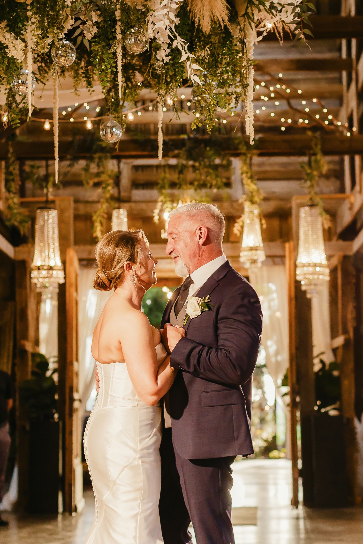 Bride and groom share first dance under hanging greenery and crystal chandeliers in elegant rustic wedding venue.