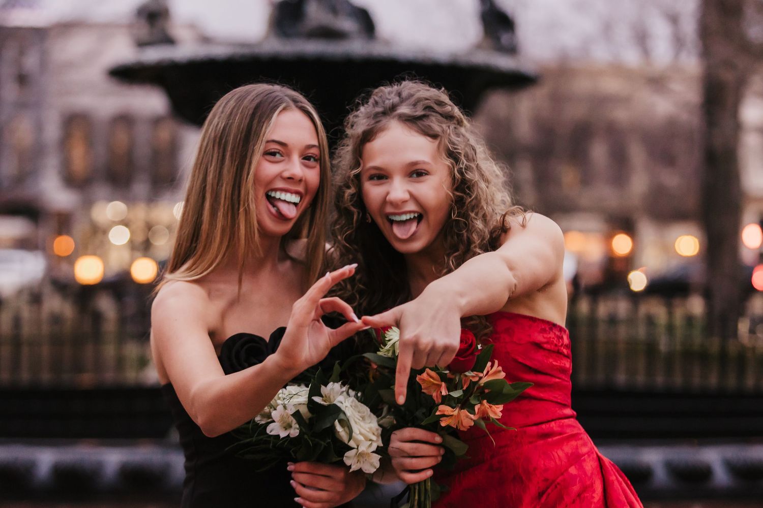 Two young women in formal dresses smile and pose with bouquets near a fountain.