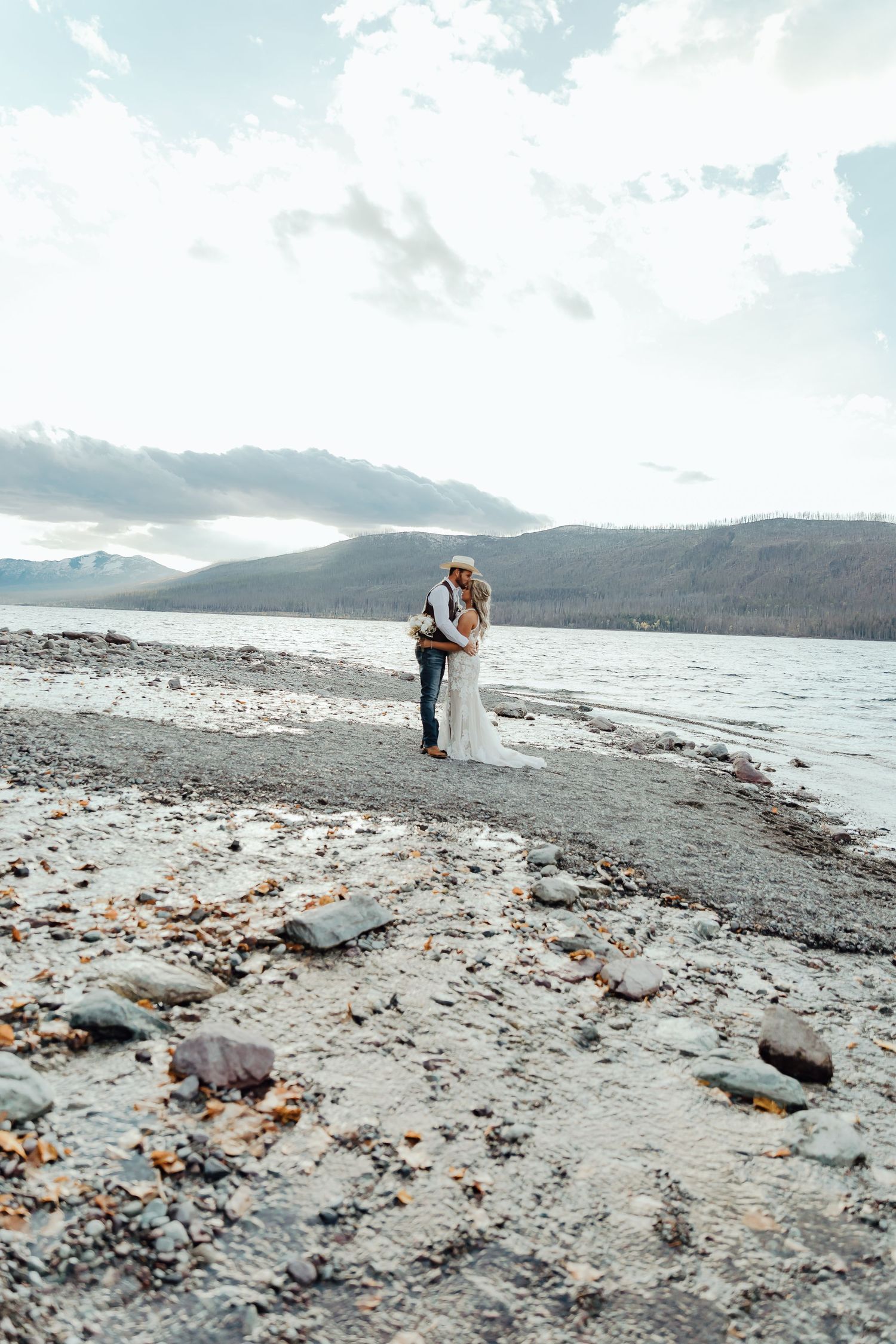 A wide angle shot of a rocky beach wedding scene with mountains and lake in the background.