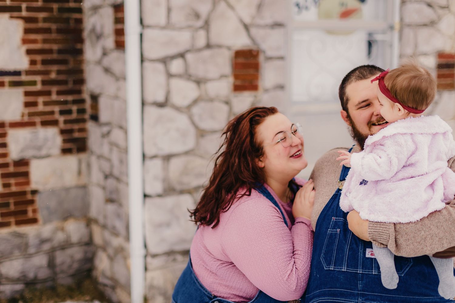 Playful family moment captured against brick and stone wall in winter.