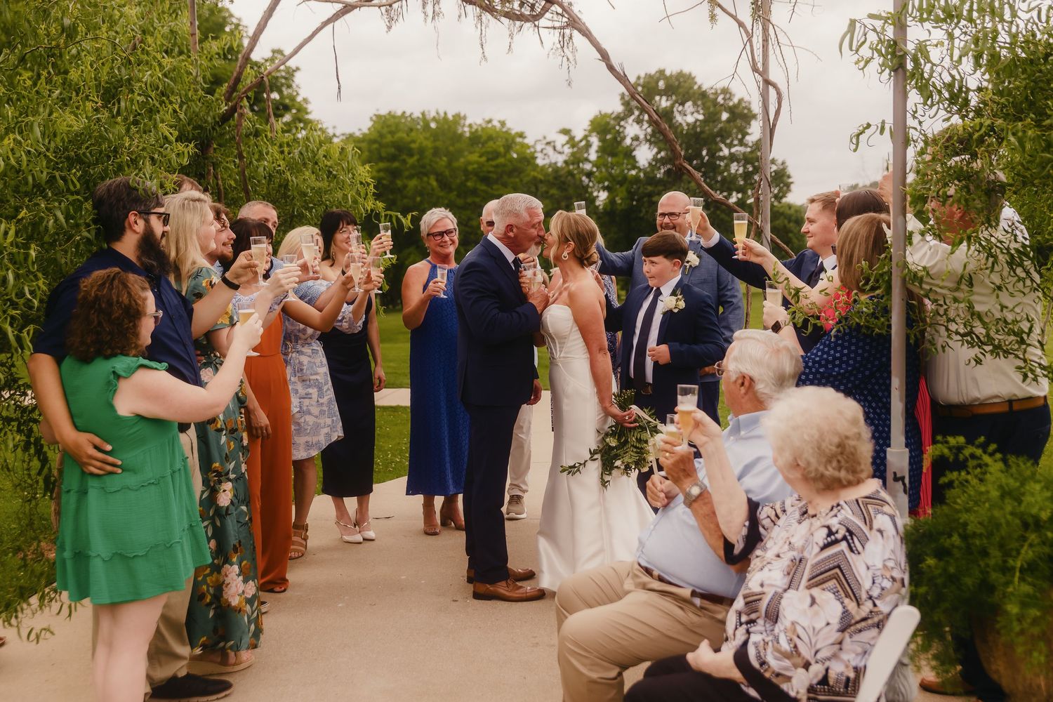 Wedding guests gather outdoors under trees during a summer reception celebration.