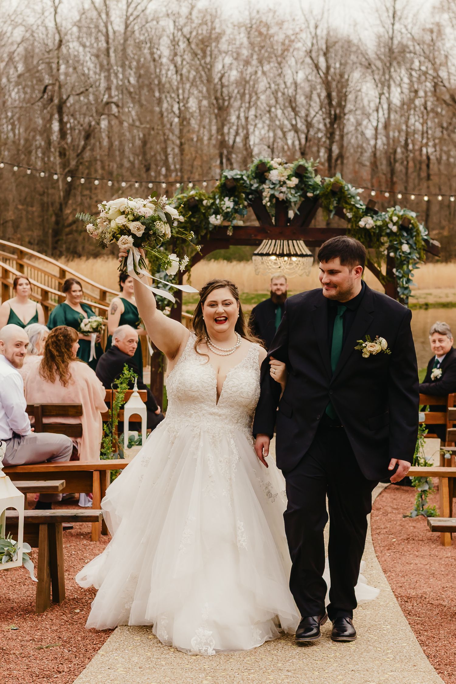 Newlyweds celebrating while walking down the aisle under floral arch at outdoor ceremony.