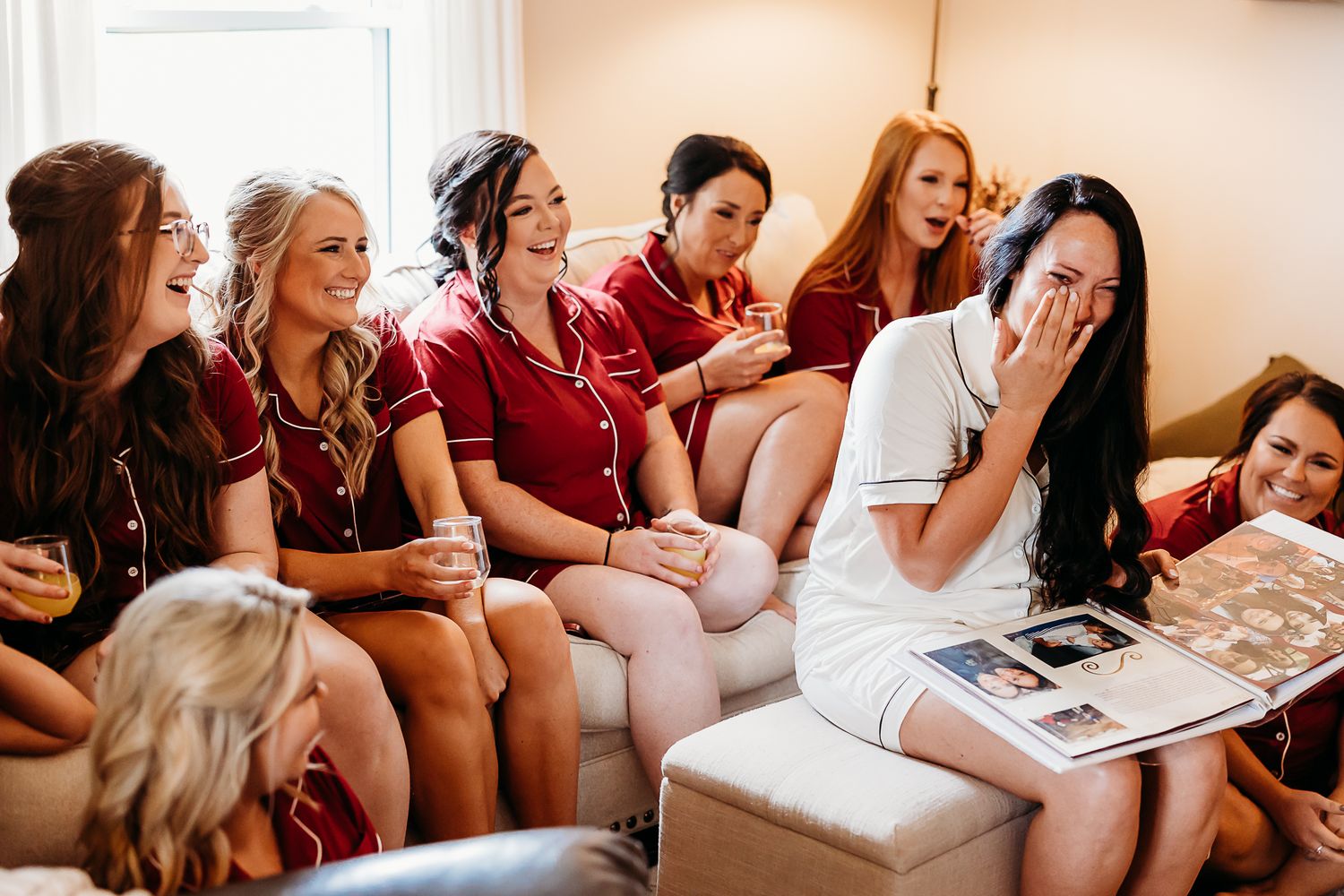 Bridesmaids in burgundy robes react emotionally while looking at photo album.