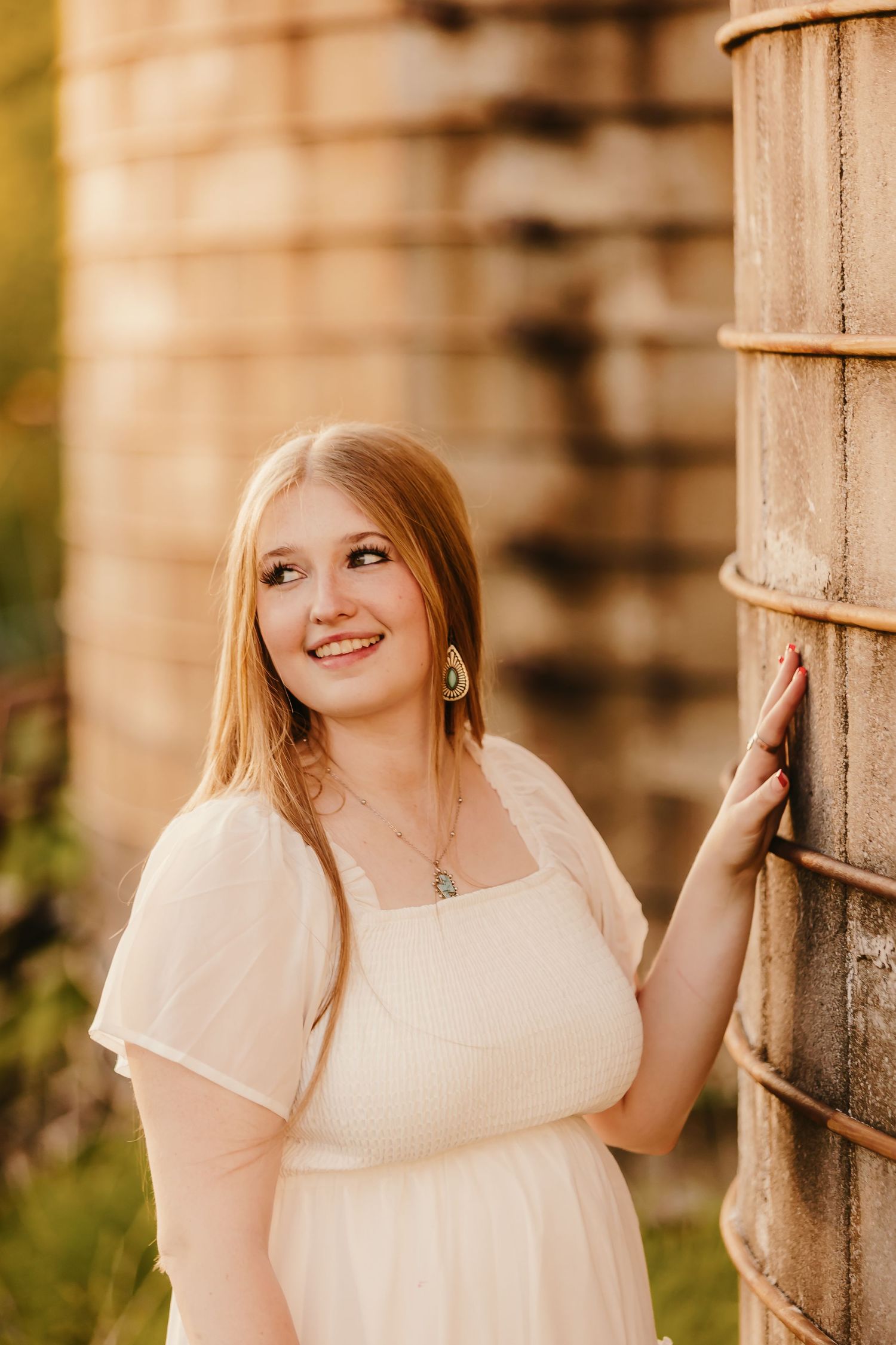 Young woman in white dress smiling beside rustic silo in golden hour light, wearing pendant necklace and earrings.