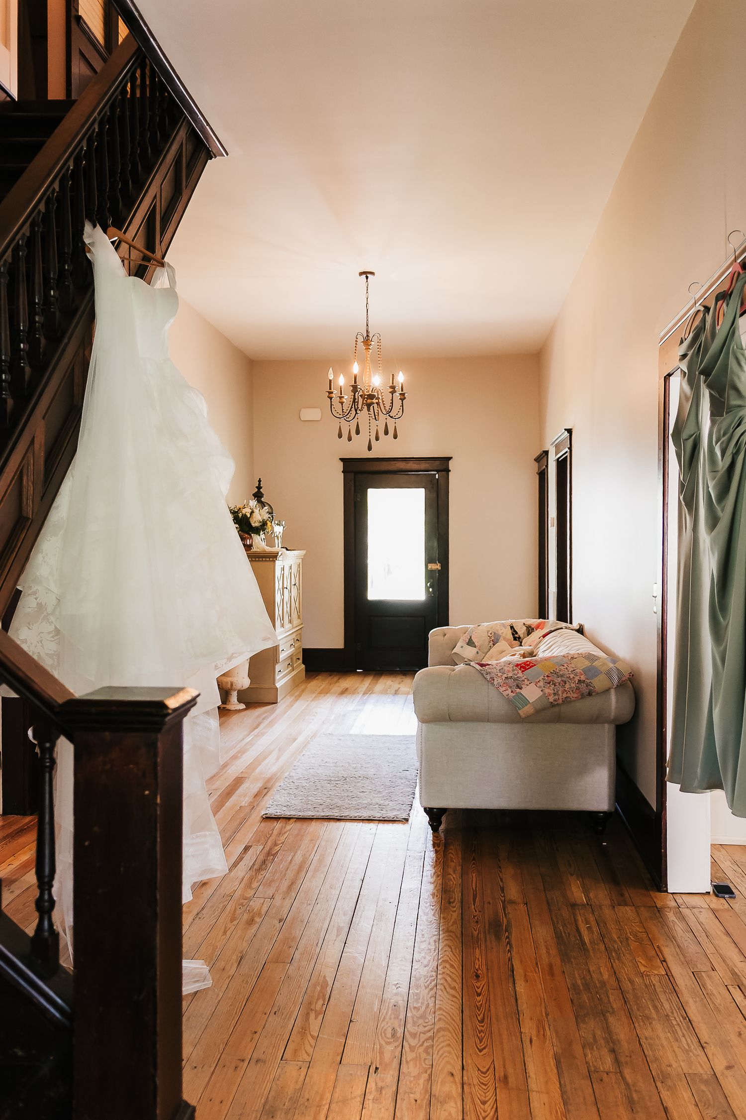 Elegant wedding venue foyer with white bridal gown hanging on staircase, chandelier, hardwood floors, and green bridesmaid dress.