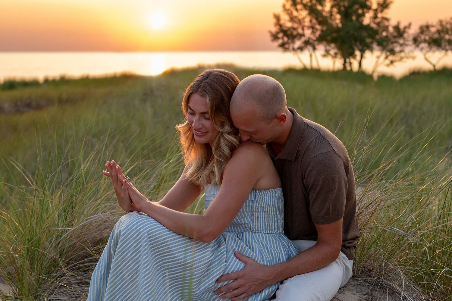 Couple sitting in grass at sunset, man kissing woman's cheek as she smiles, ocean visible in background.