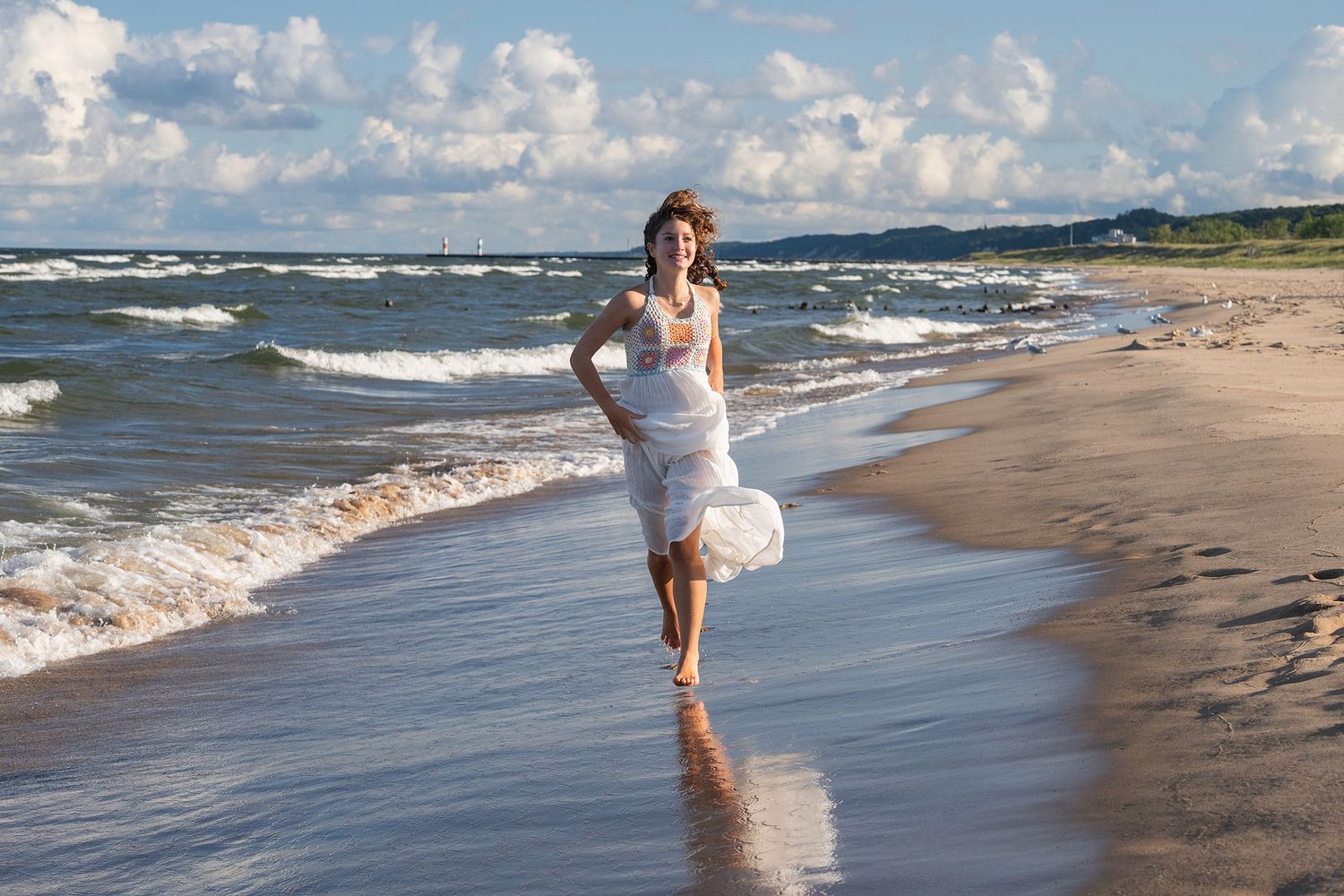 A person in white runs joyfully through the surf along a scenic beach with rolling waves.