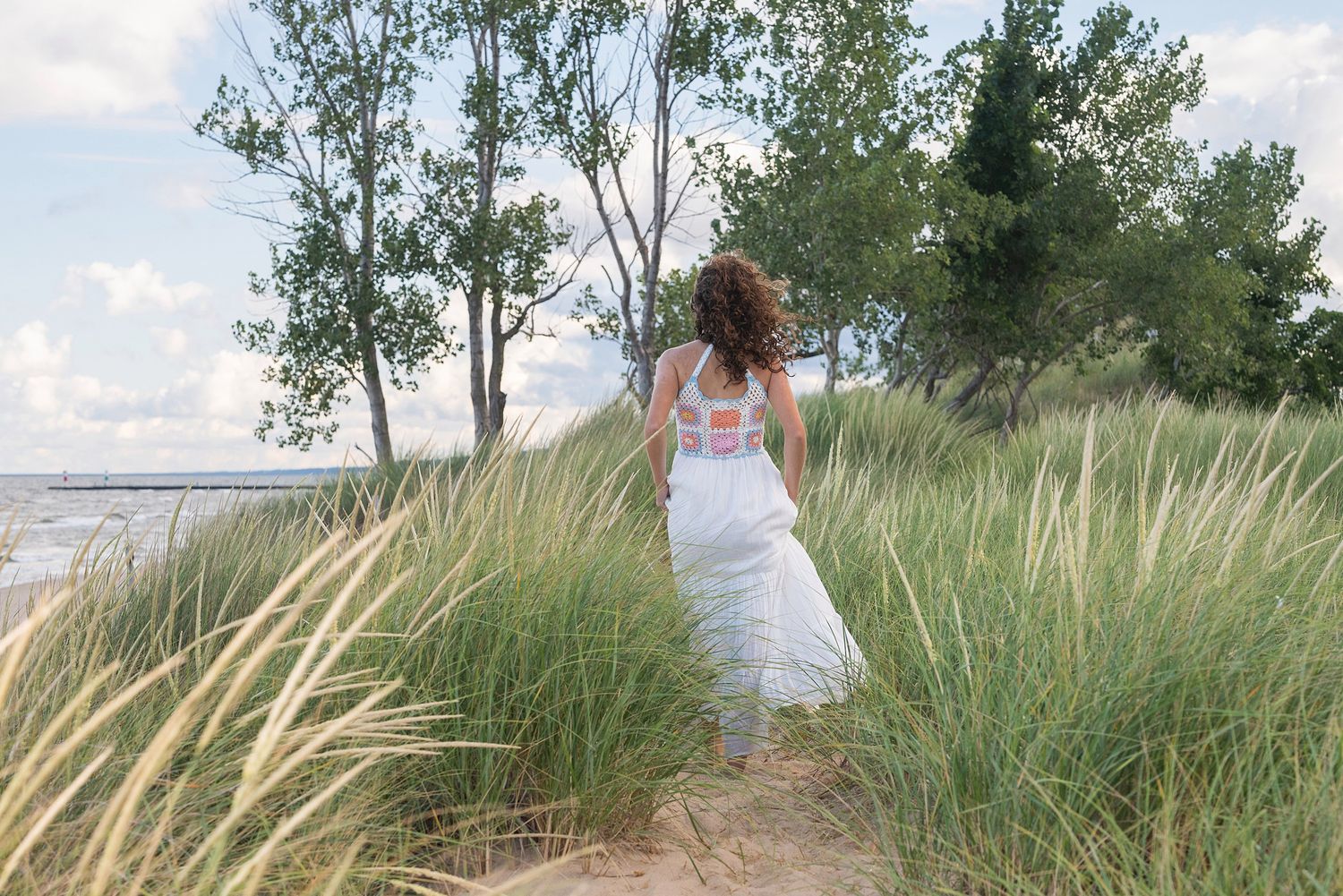 Figure in white walking through windswept dune grass against a bright blue sky.