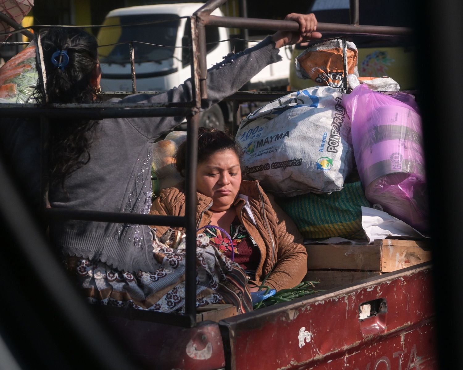 People riding in the back of a red pickup truck through a rural area.