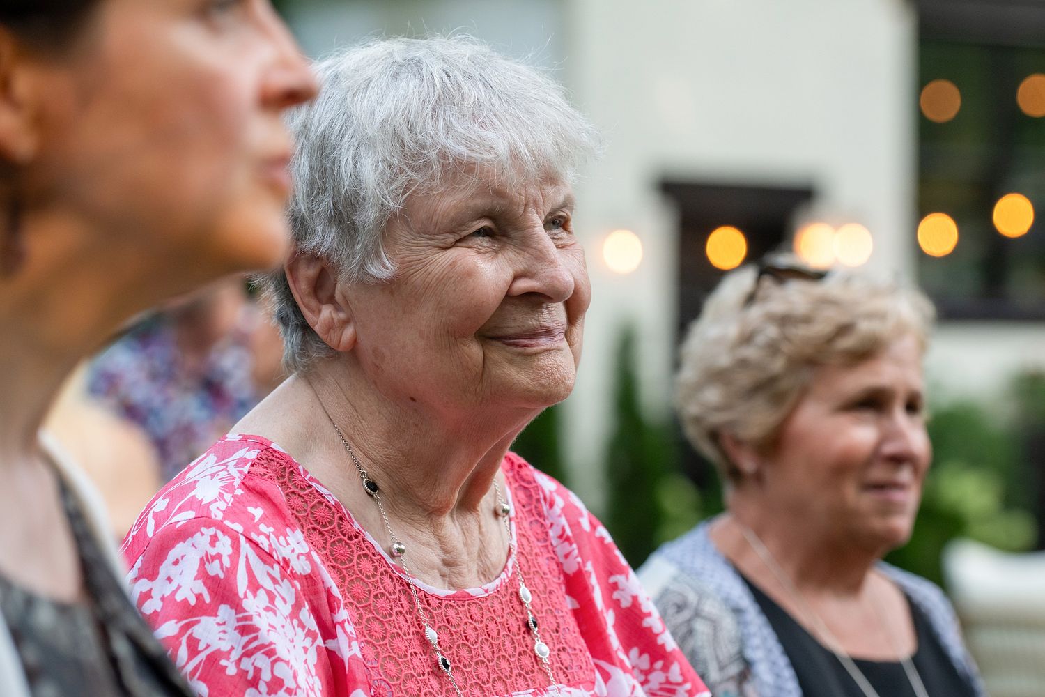 A senior woman in a bright pink blouse smiles warmly while attending an outdoor gathering.
