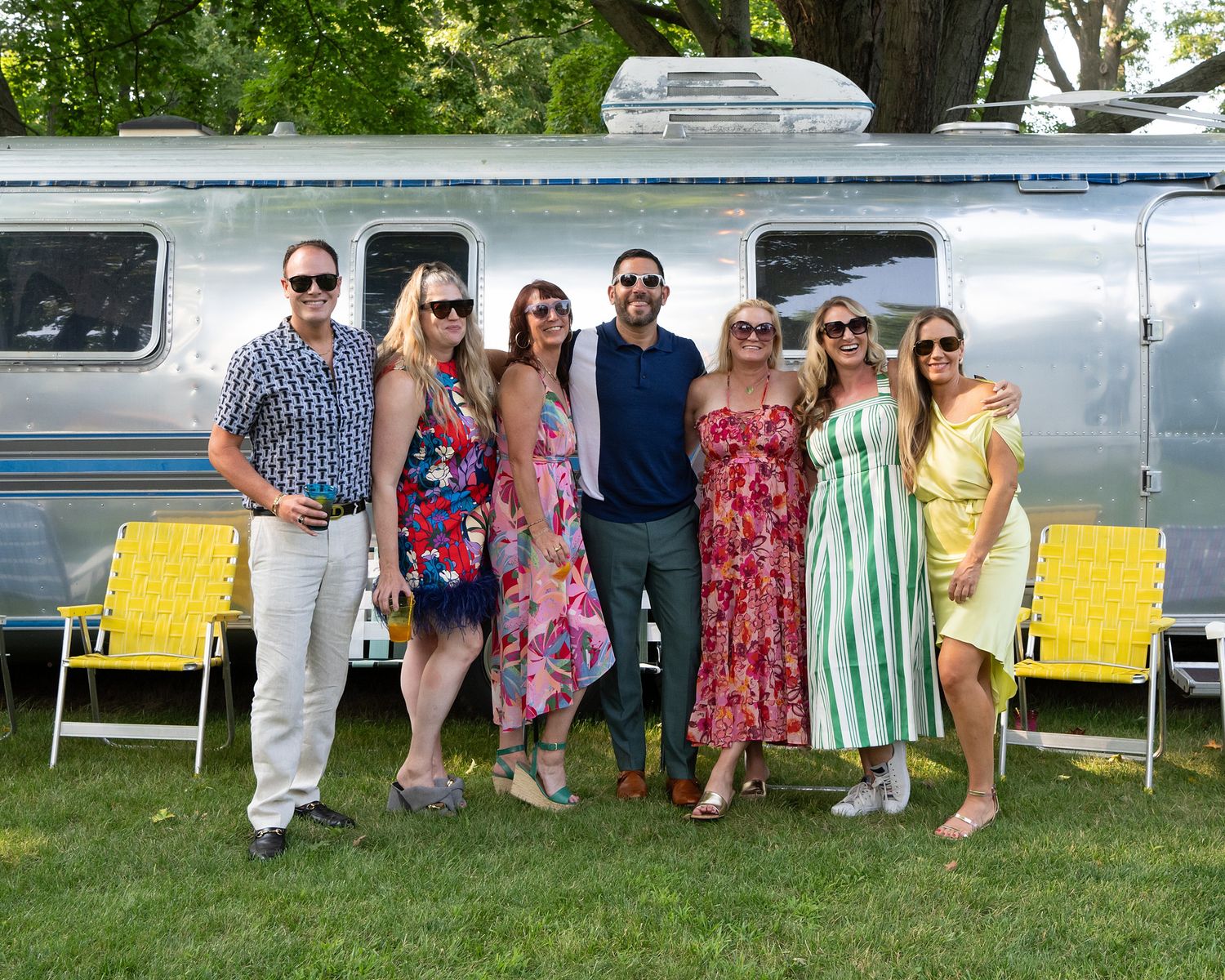 A group stands together in front of a silver Airstream trailer with yellow lawn chairs on a sunny summer day.