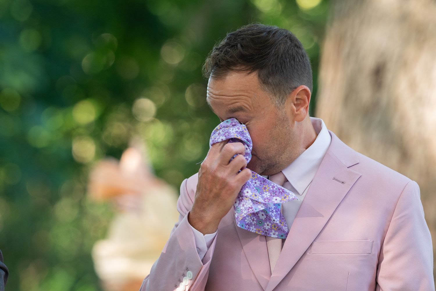 Someone in a pink suit jacket wipes tears with a purple handkerchief during an emotional outdoor moment.