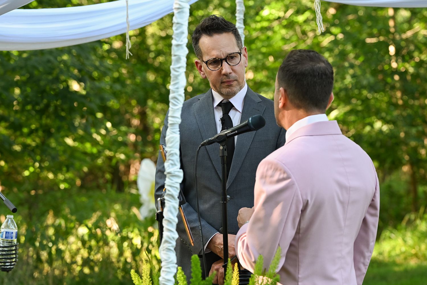A couple exchanges vows during an outdoor wedding ceremony beneath white fabric draping and green trees.
