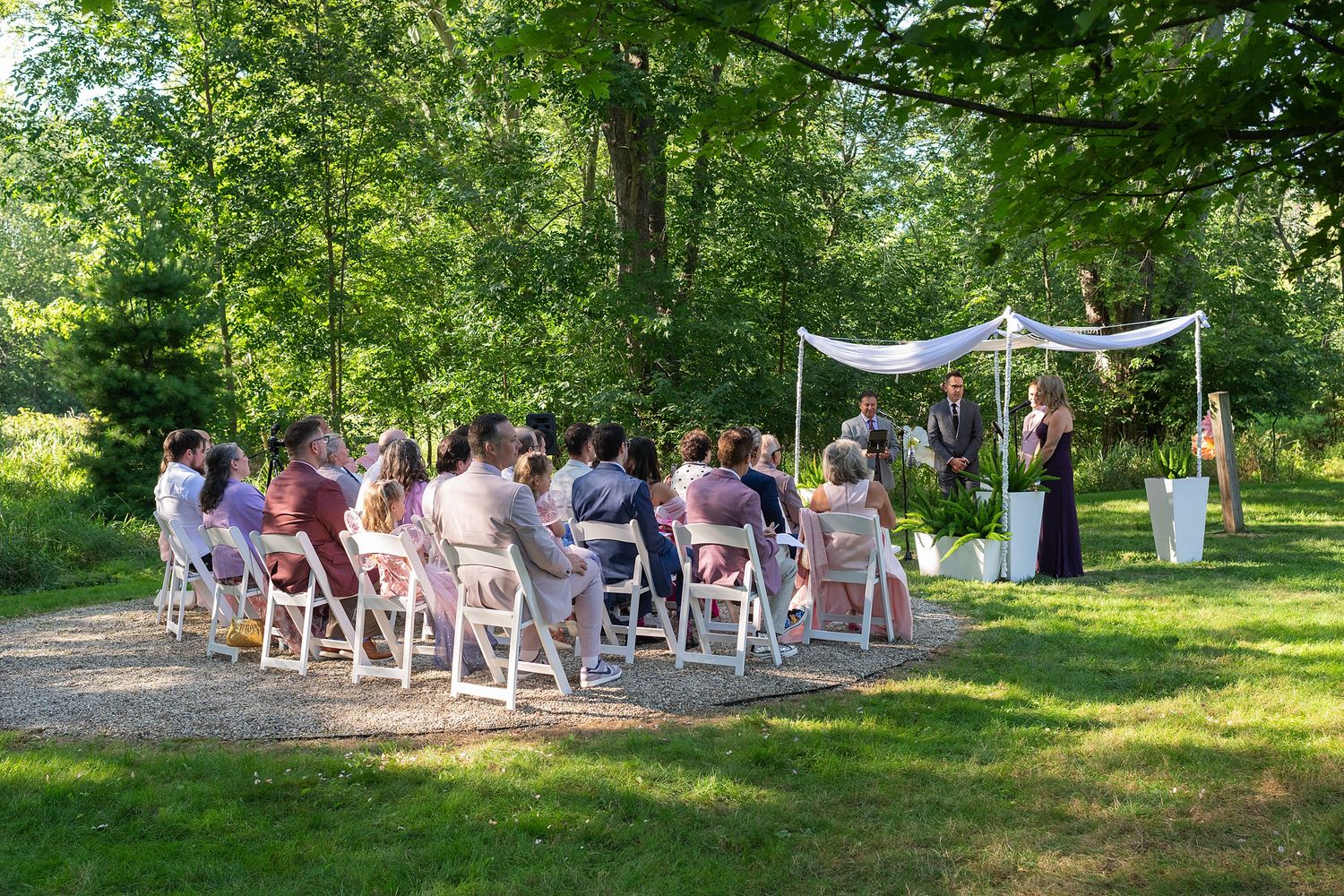 White folding chairs arranged outdoors for a garden ceremony under green trees on a sunny summer day.