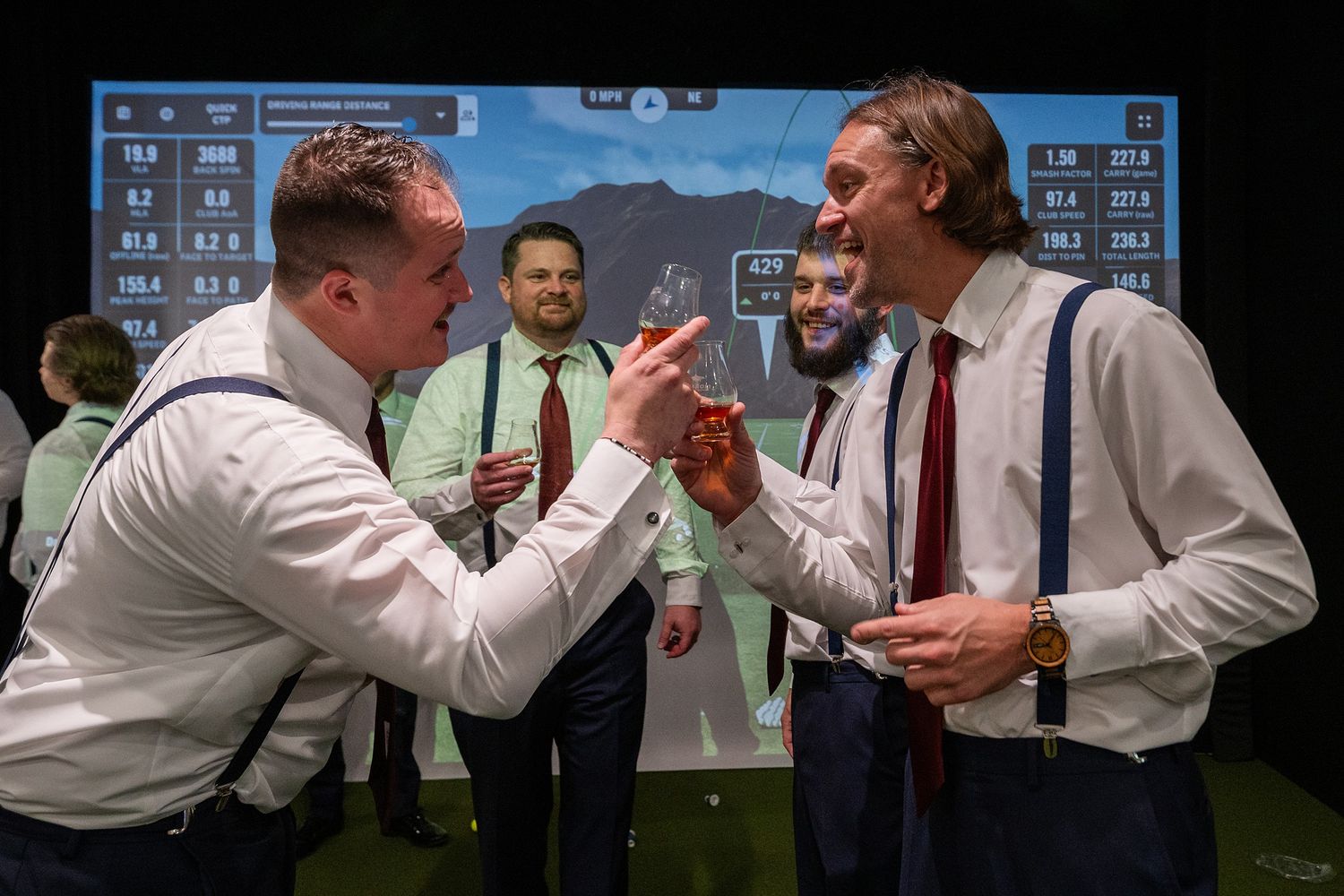 Two men in dress shirts and suspenders laugh together at an indoor golf simulator event.