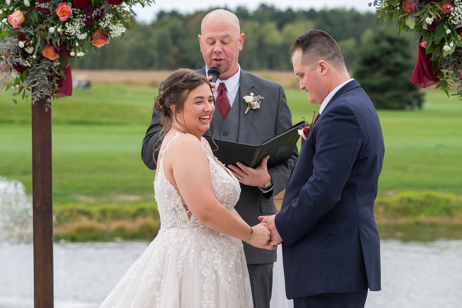 Bride and groom exchange vows outdoors at wedding ceremony with officiant, floral arch, and scenic pond backdrop.