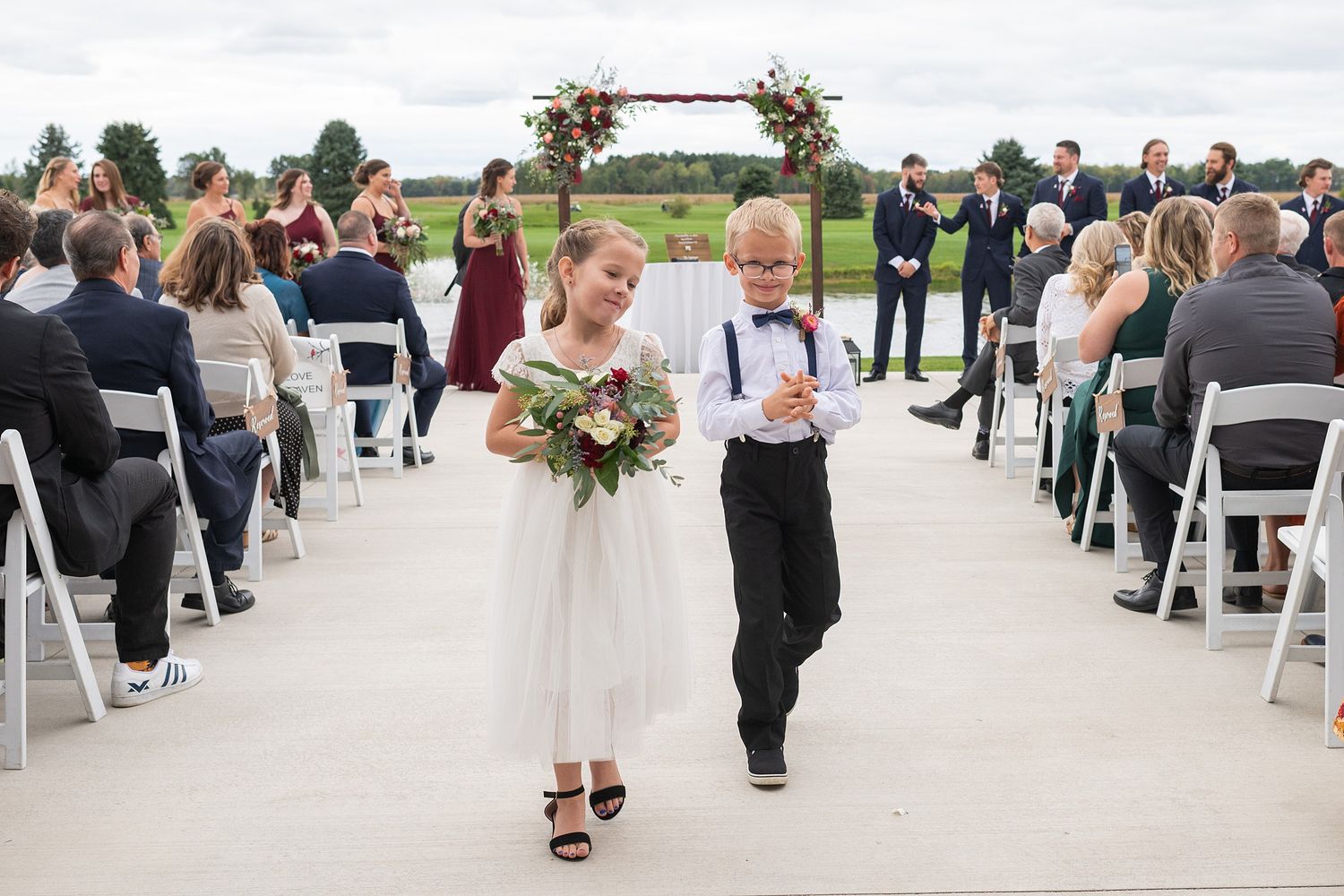 Young flower girl and ring bearer walk down wedding aisle at outdoor ceremony.