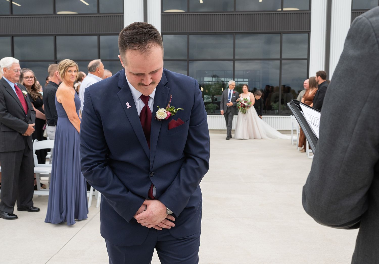 Emotional groom in navy suit bowing his head as bride walks down the aisle at outdoor ceremony.