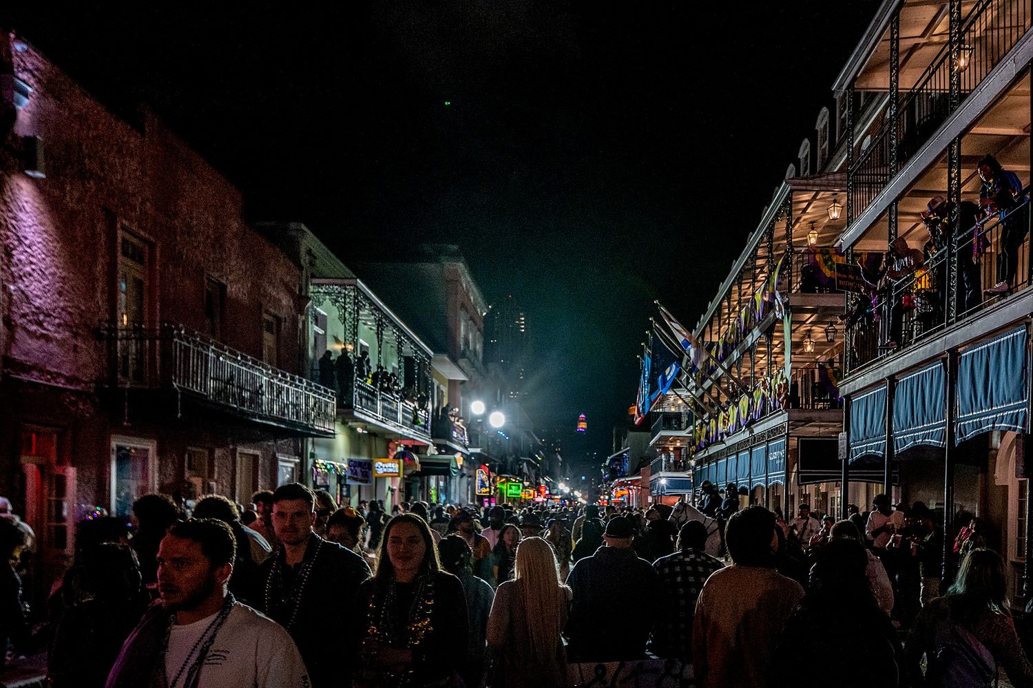 Nighttime panoramic view of crowded Bourbon Street with illuminated buildings and neon signs.