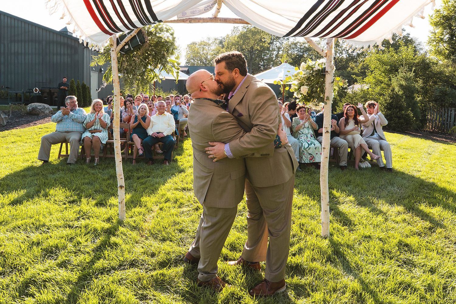 Two grooms in tan suits embrace under a striped wedding canopy on a sunny lawn as seated guests watch the outdoor ceremony.