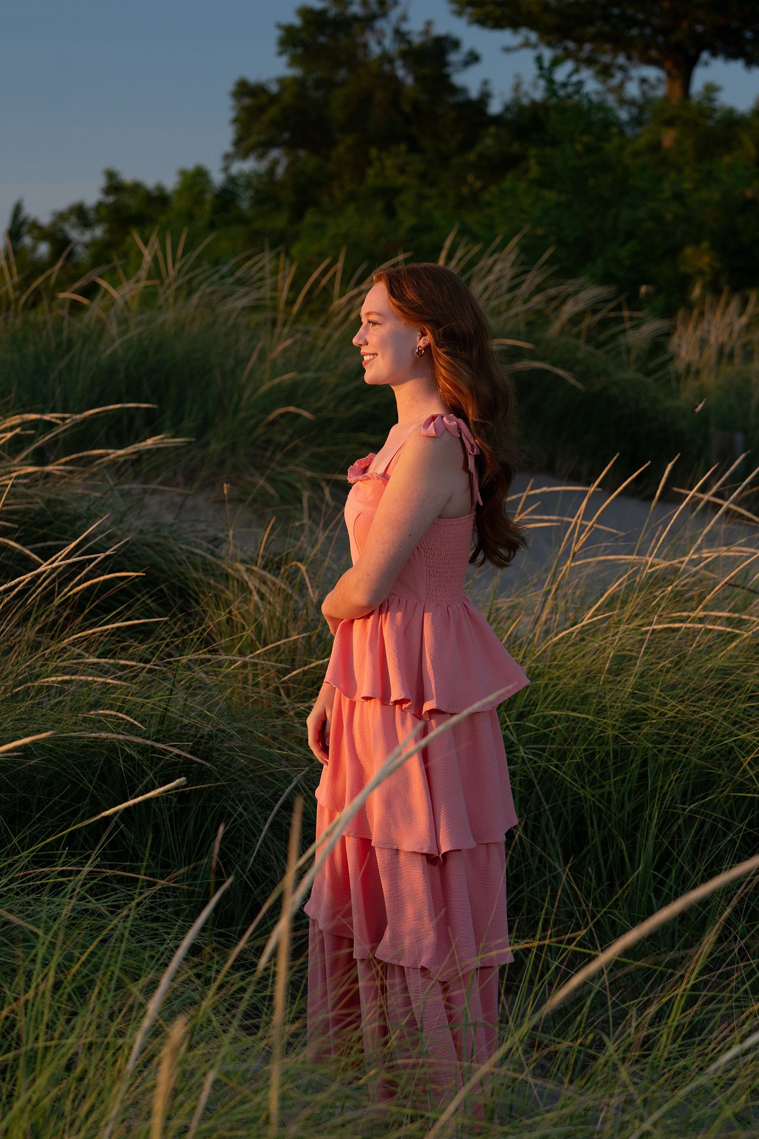 Young woman in pink ruffled maxi dress standing among tall grass at golden hour sunset.