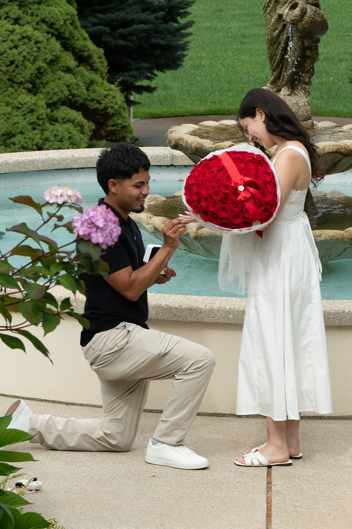 Man kneeling on one knee presenting a ring and red roses bouquet to woman in white dress by a garden fountain.