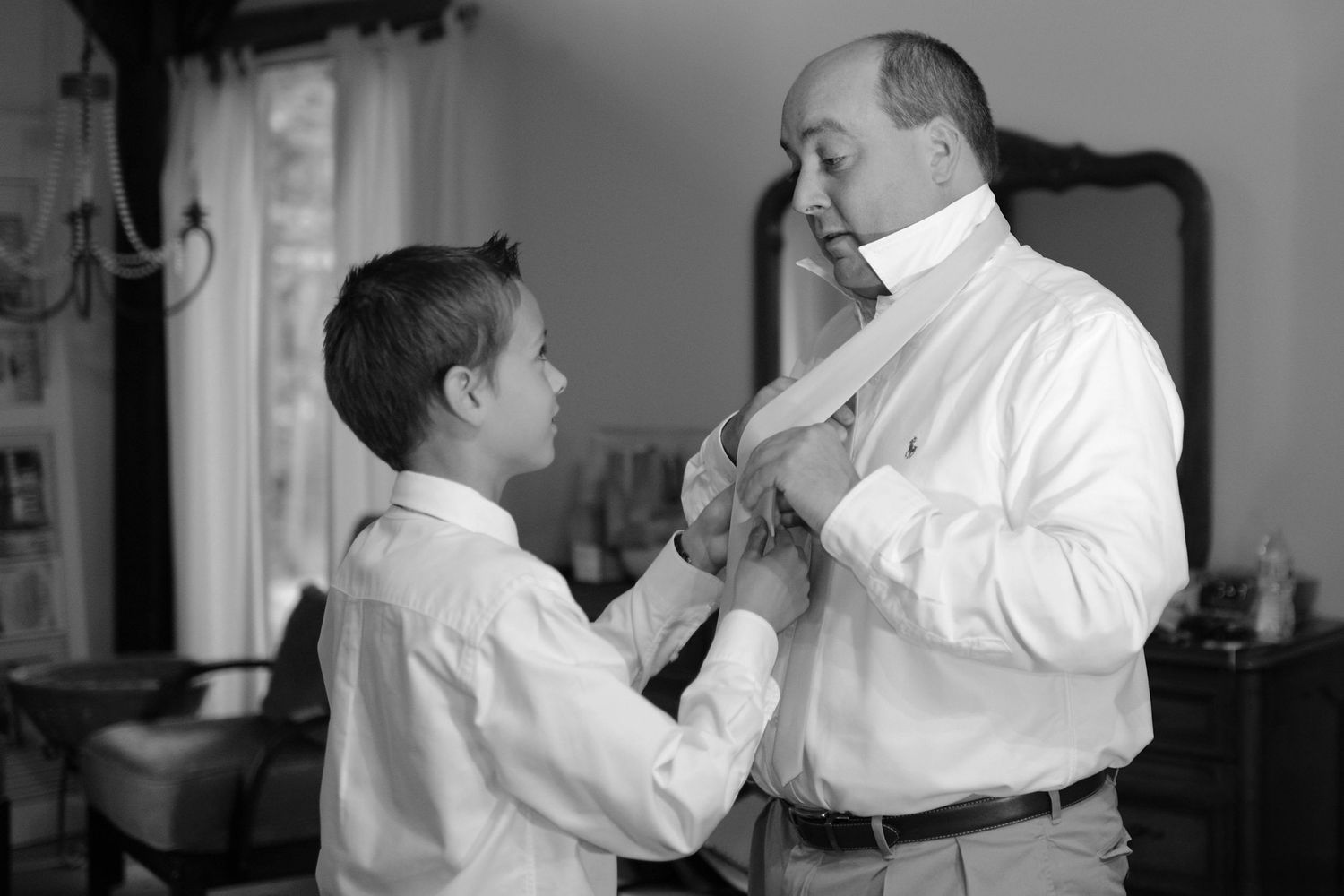 Black and white photo of a father and son in dress shirts preparing for a formal event together.